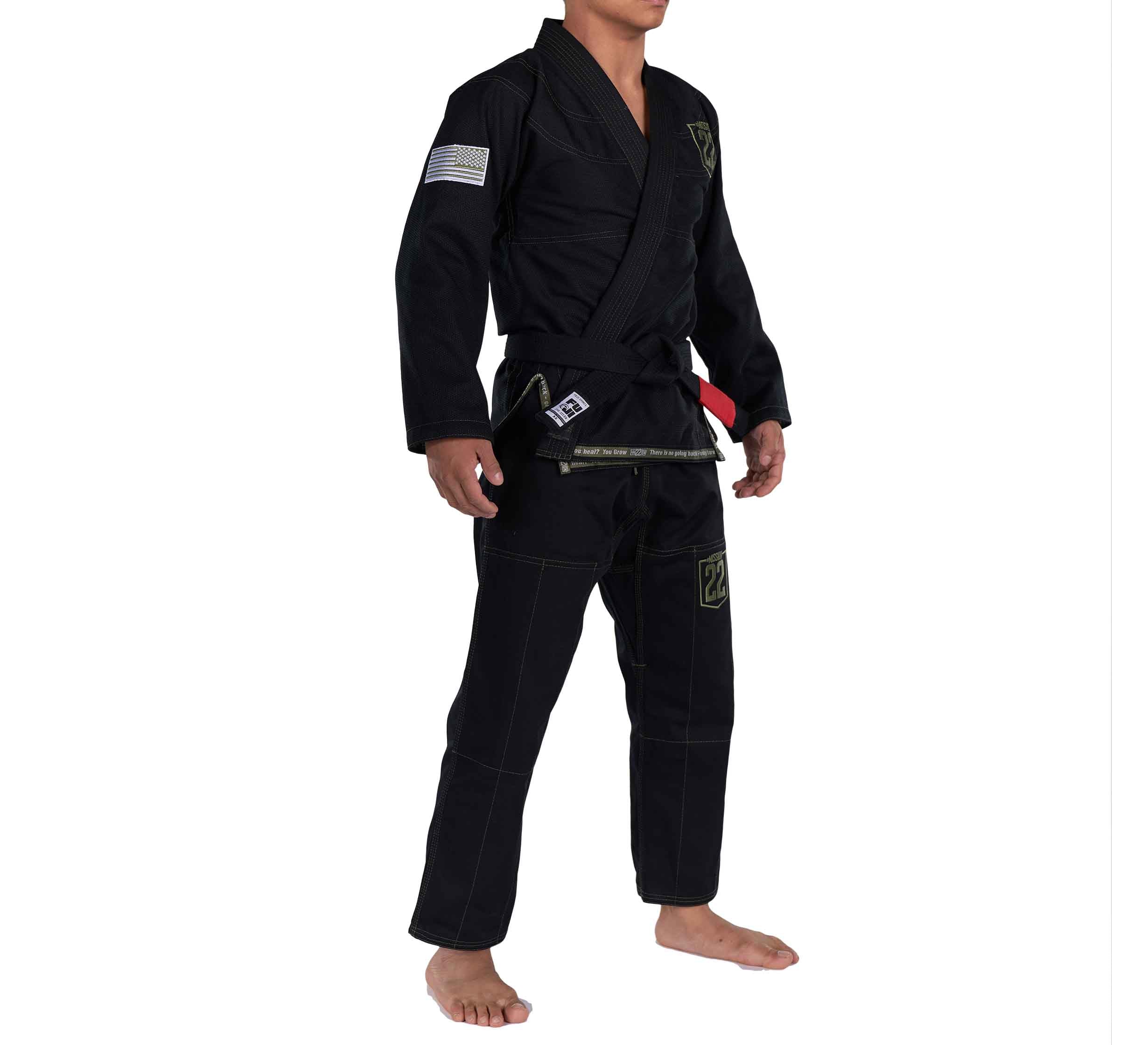 A person stands barefoot against a white background wearing the Mission 22 Black Gi with a black belt. The gi features patches, including an American flag on the sleeve.