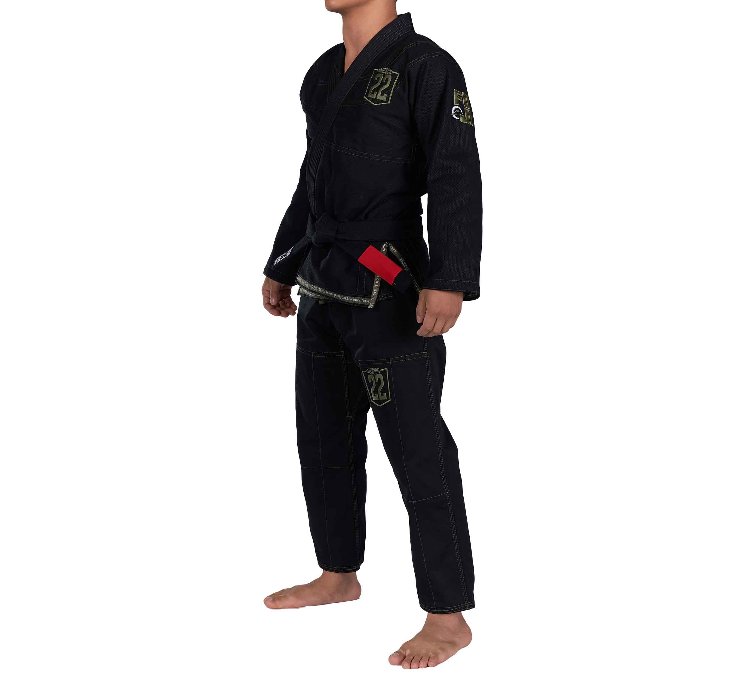 A person stands barefoot on a white background wearing the Mission 22 Black Gi, which includes a jacket, pants, and a black belt with a red tag. The gi features patches, and the head is cropped out of the image.