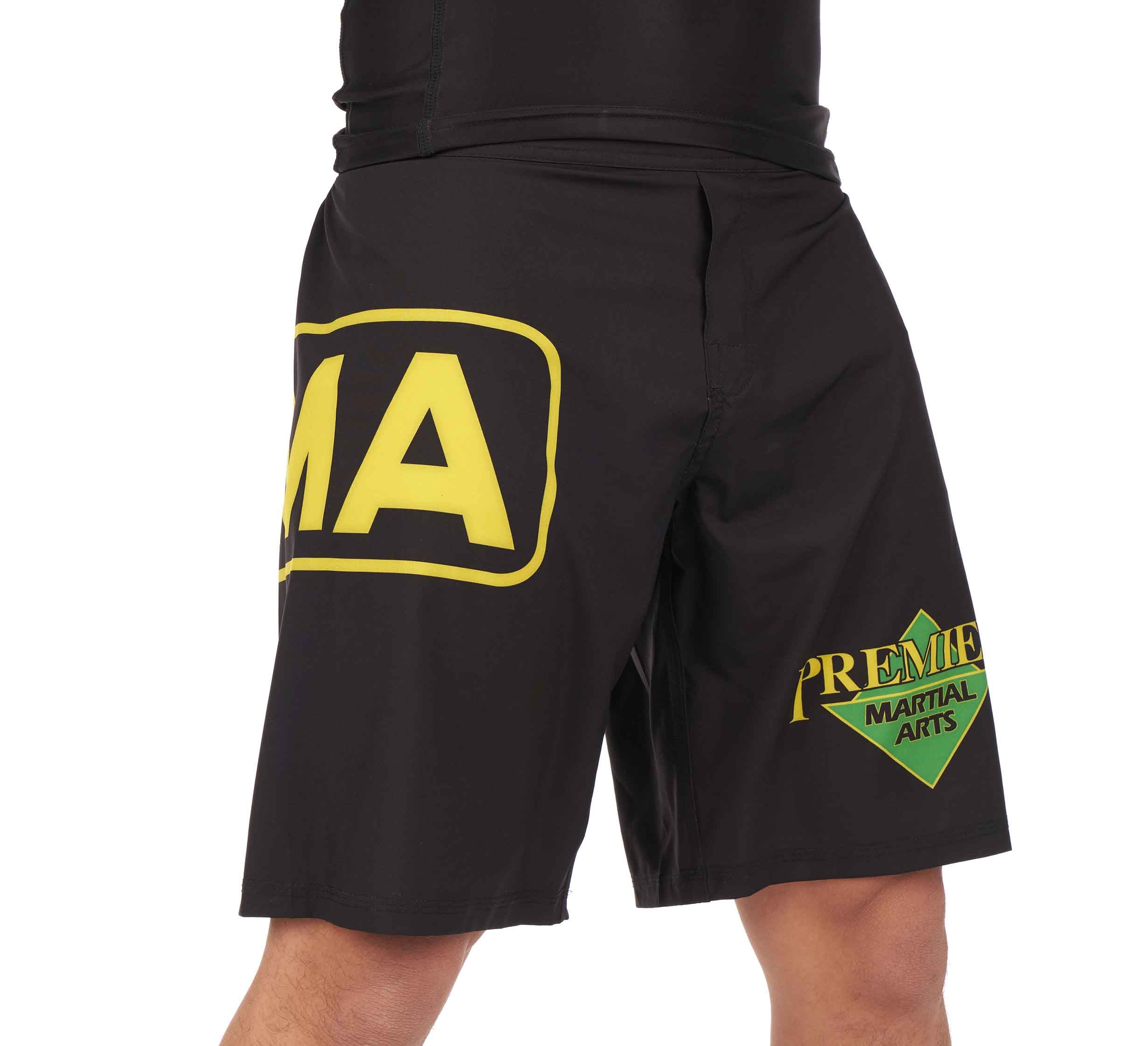 Someone wearing Premier Martial Arts Fight Shorts, which are black with yellow and green PREMIER MARTIAL ARTS logos on the left leg and a partial MA logo on the right leg.