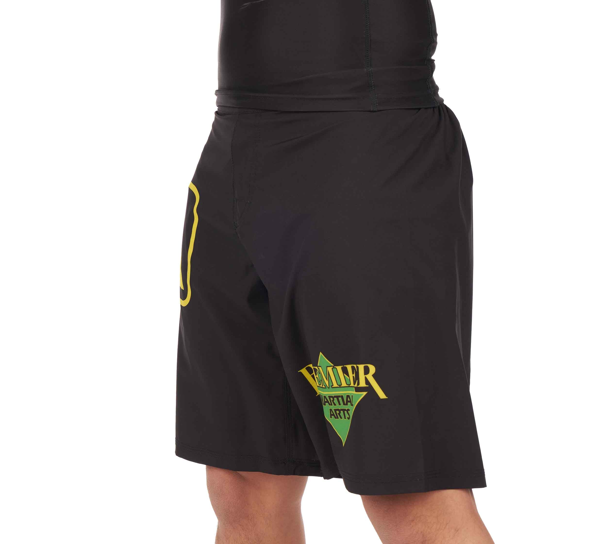 A person wearing Premier Martial Arts Fight Shorts, which are black with yellow and green lettering and graphics on the left leg; only the lower torso and upper legs are visible.