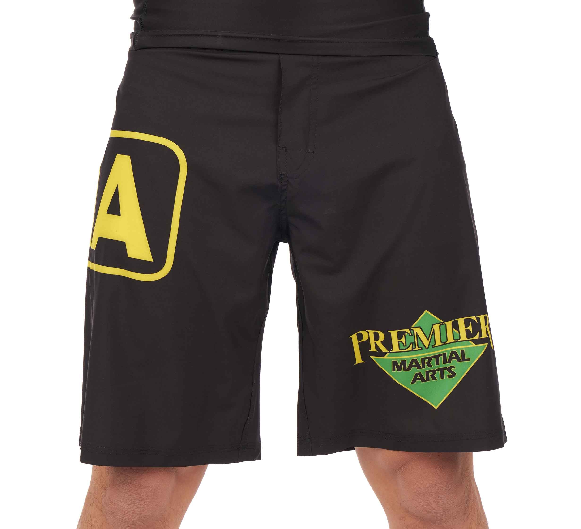 A person’s lower torso and upper legs are shown wearing Premier Martial Arts Fight Shorts, black with a large yellow A on the left thigh and a green and yellow Premier Martial Arts logo on the right thigh.