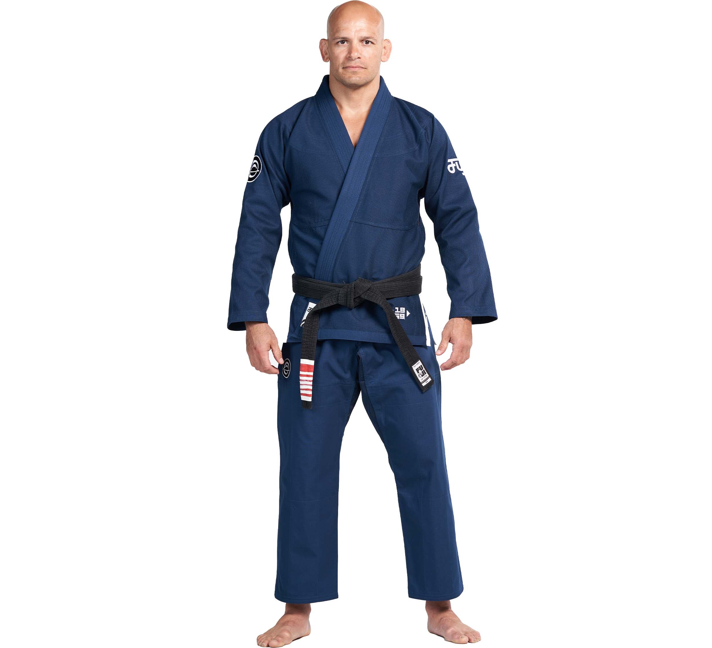 A man stands barefoot, facing forward with a neutral expression, wearing the Flow-Tech BJJ Gi Navy with a black belt. The gi features white patches and logos on the arms and legs, set against a plain white background.