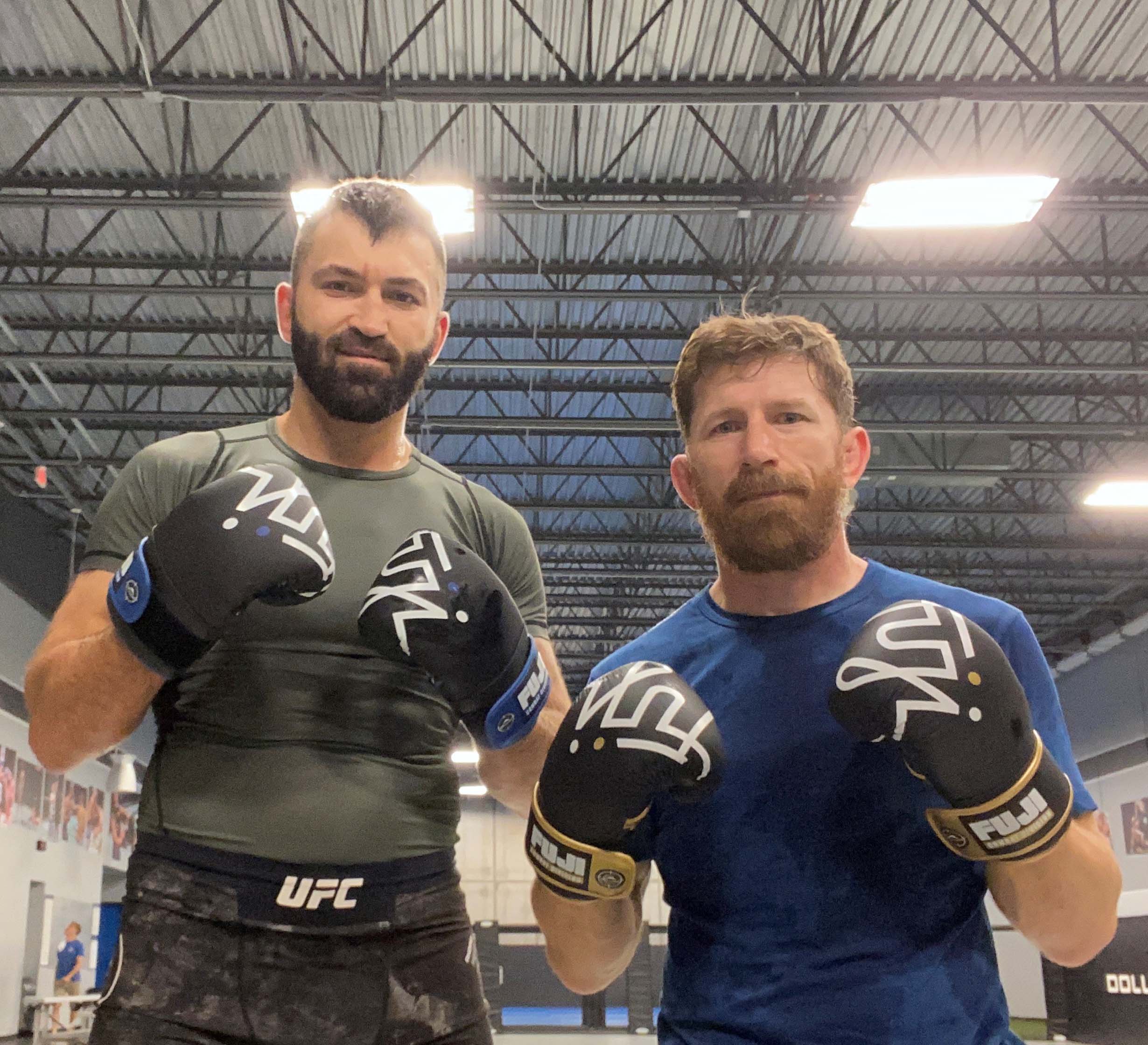Two men pose in a gym under bright lights, both wearing FUJI Ultimate Hybrid Gloves. One sports a dark athletic shirt and camo shorts, the other wears blue. They face the camera, ready to spar among metal beams.