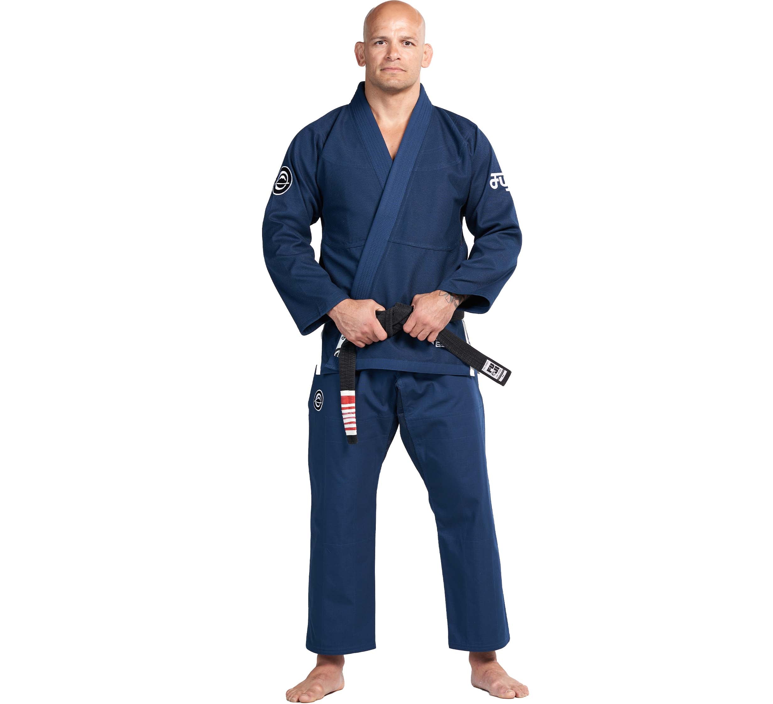 A man stands barefoot in a Flow-Tech BJJ Gi Navy, holding a black belt with red stripes. He has a serious expression and poses against a plain white background.