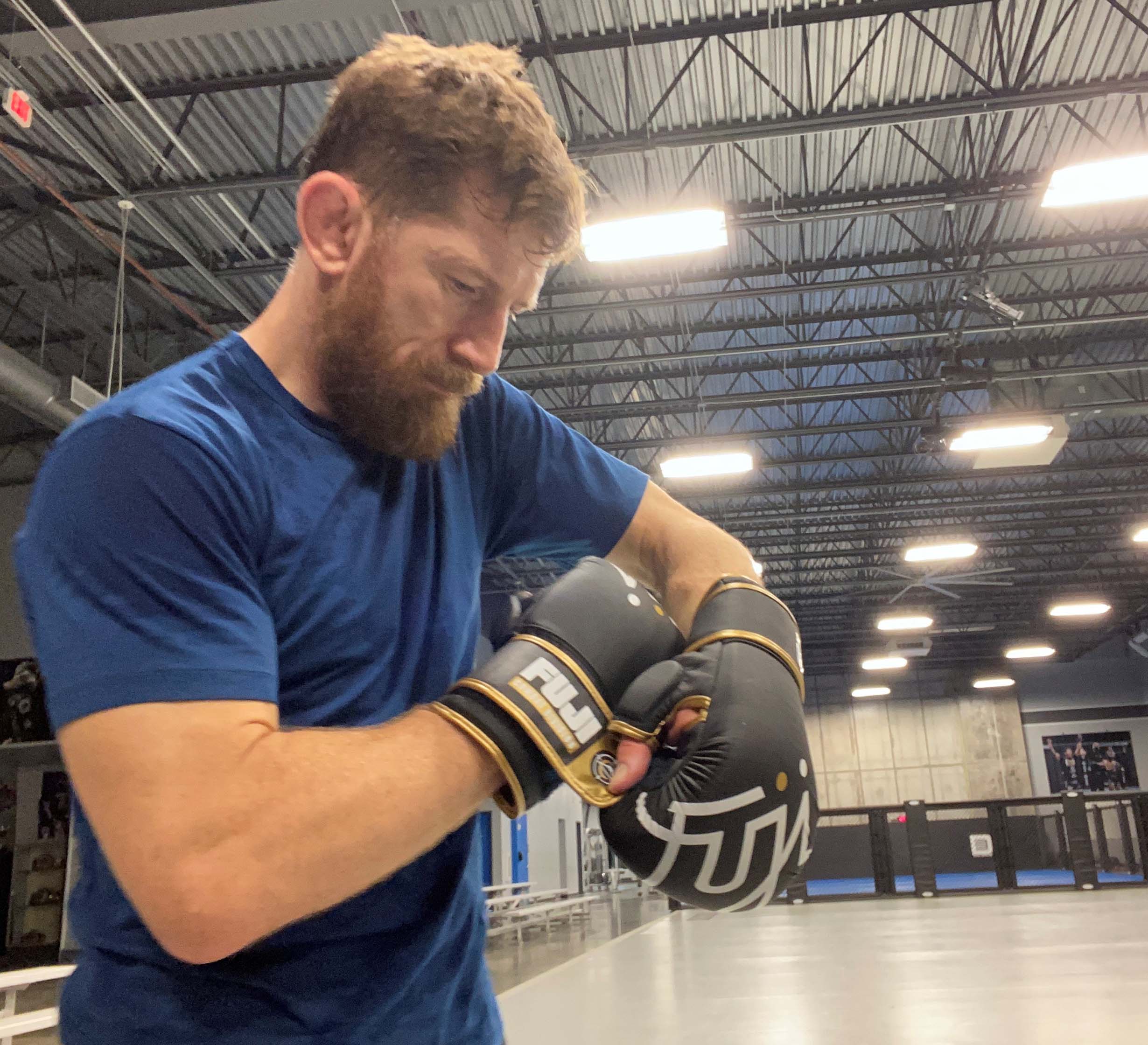 A bearded man in a blue shirt stands in a brightly lit gym with high ceilings, looking down as he adjusts his black FUJI Ultimate Hybrid Gloves in the open training area.