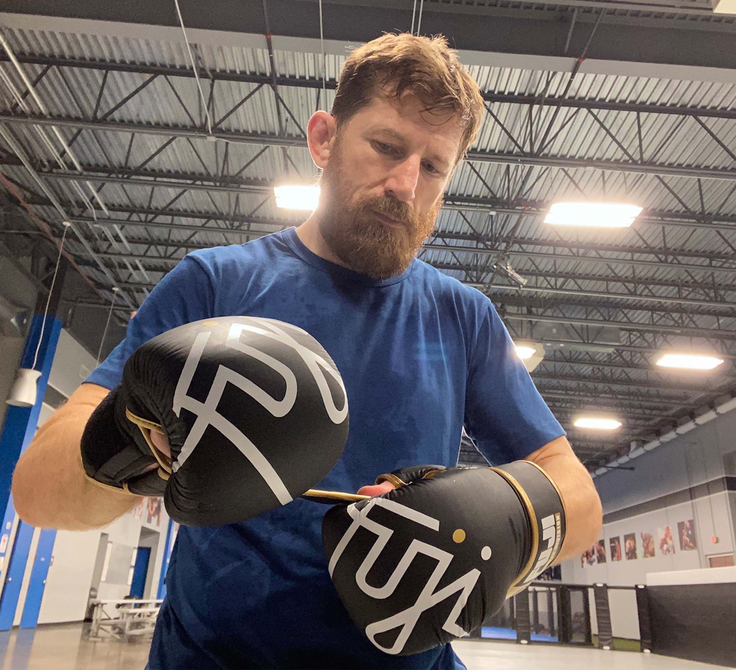 A bearded man in a blue shirt tightens the strap on his FUJI Ultimate Hybrid Gloves inside a spacious, well-lit gym with high ceilings and visible equipment in the background.