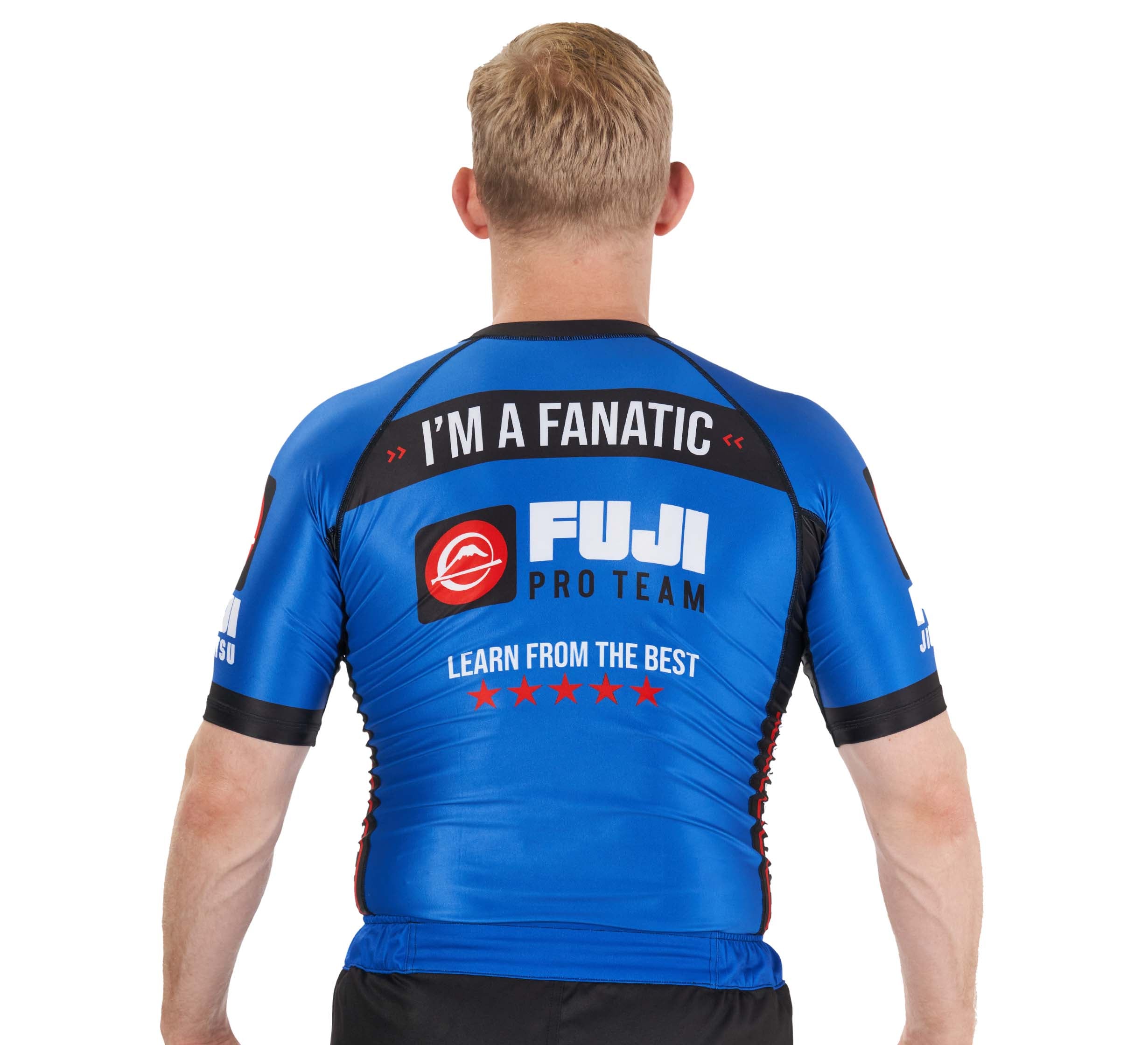 A person with short blond hair stands with their back to the camera, wearing a tight blue BJJ Fanatics Rashguard featuring "IM A FANATIC" and "LEARN FROM THE BEST" on the back.