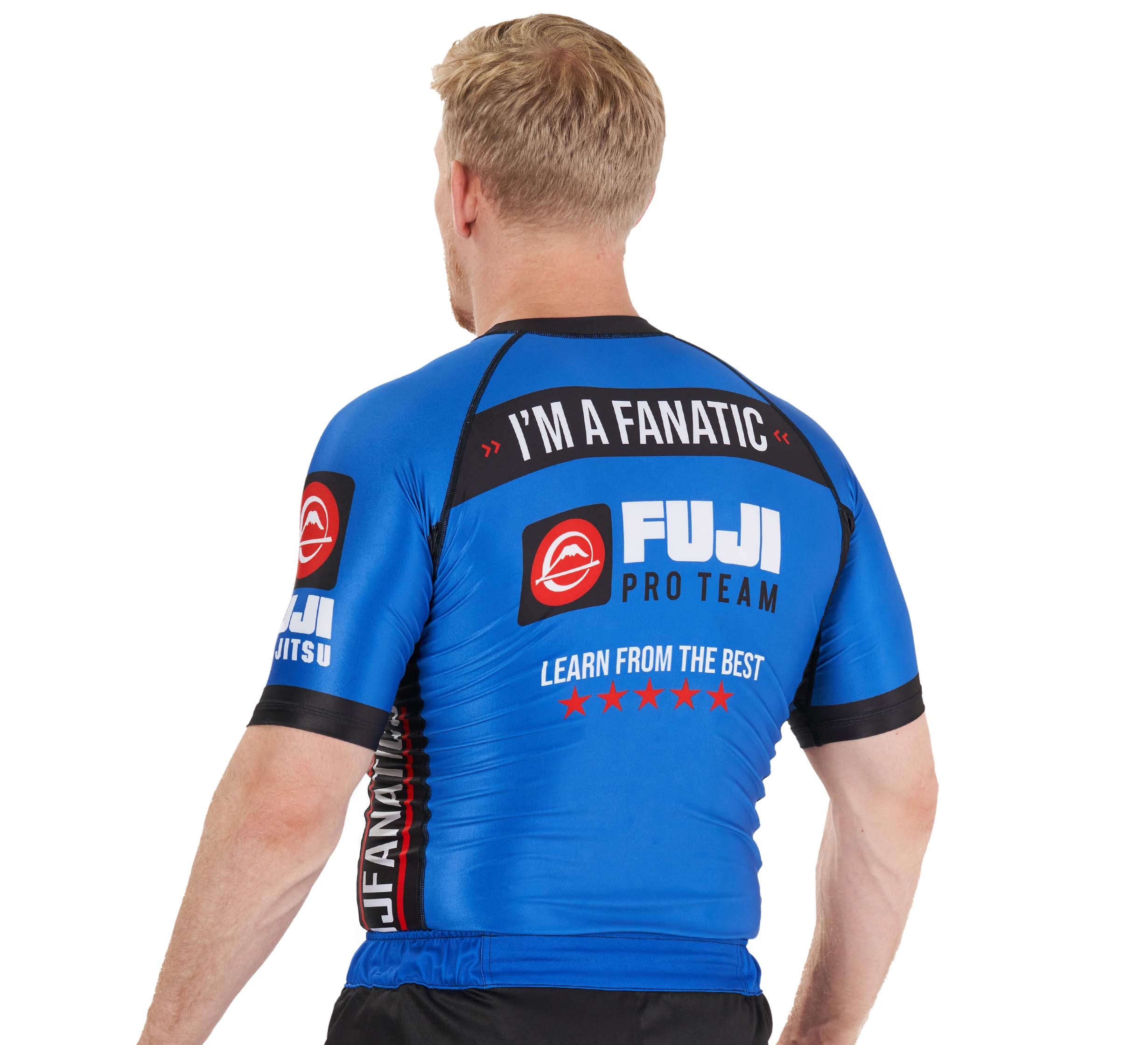 A person with blond hair is wearing a blue BJJ Fanatics Rashguard. The back of the shirt reads: "I'M A FANATIC," "BJJ Fanatics," and "LEARN FROM THE BEST.