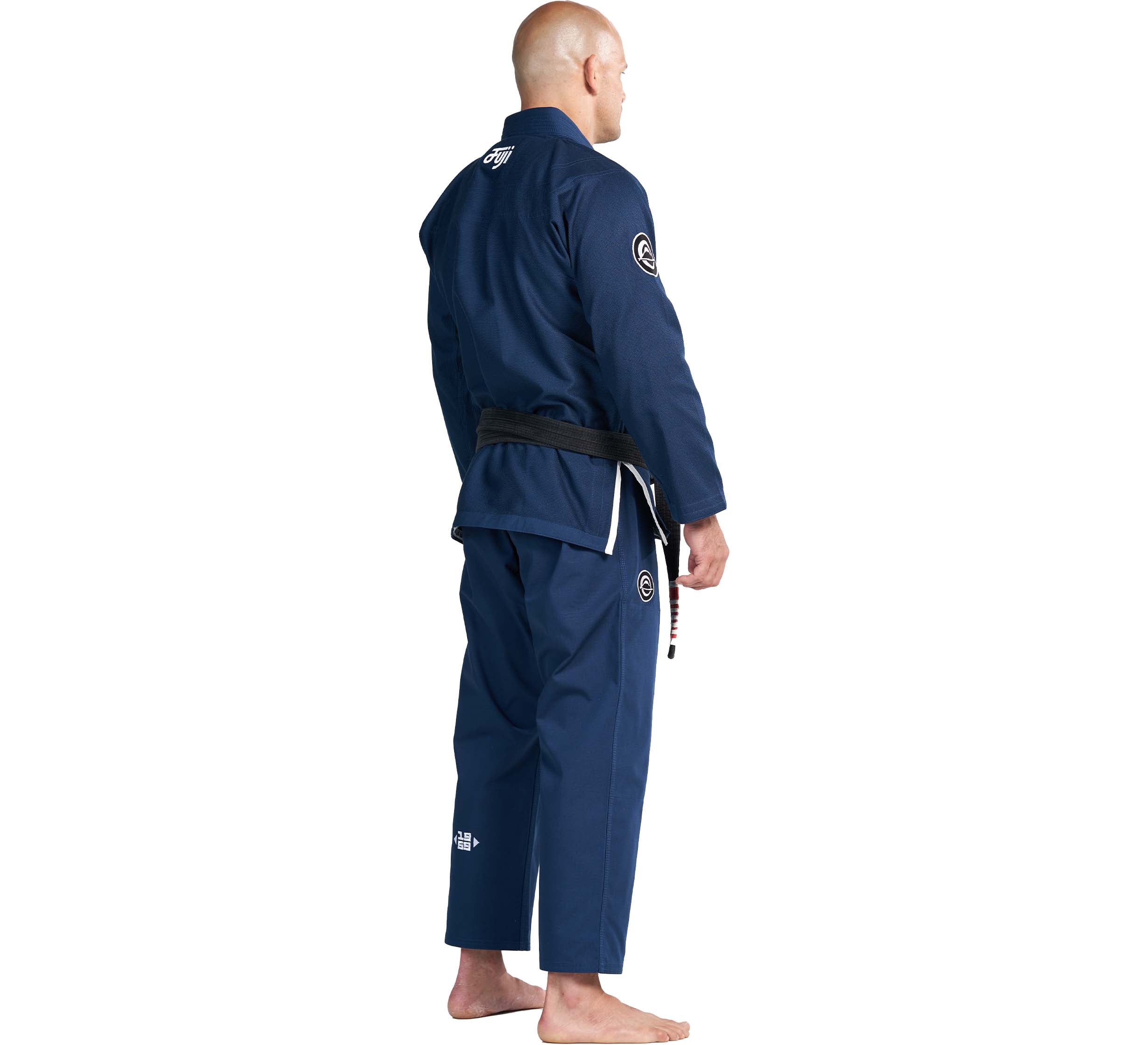 A barefoot man in the Flow-Tech BJJ Gi Navy with a black belt stands facing away, looking to the side. His head is shaved and patches are visible on the jacket and pants.
