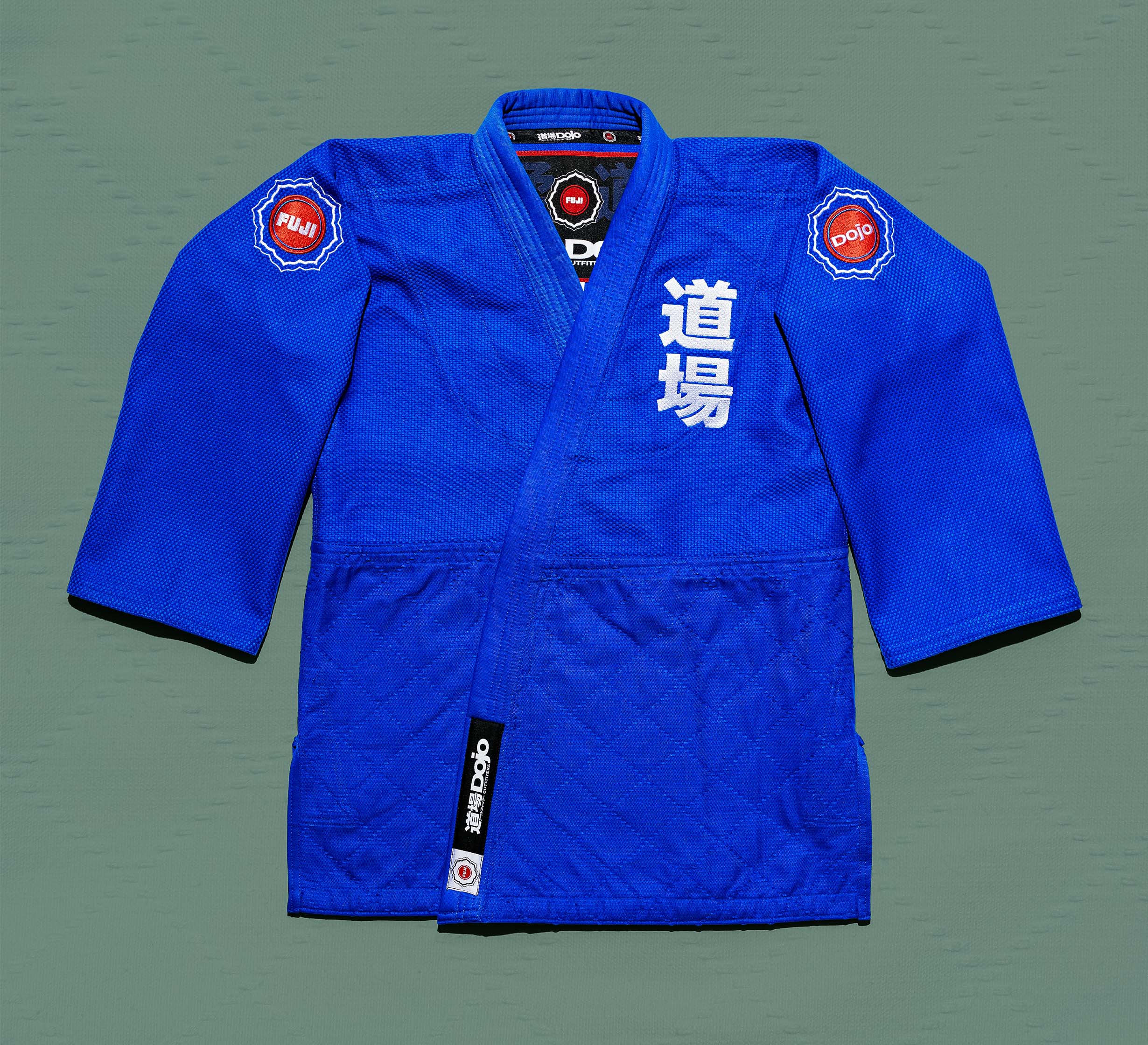 A Dojo Outfitters x FUJI Judo Gi Blue jacket featuring Japanese characters on the left and Fuji/Dojo patches on the shoulders, shown flat against a green background.