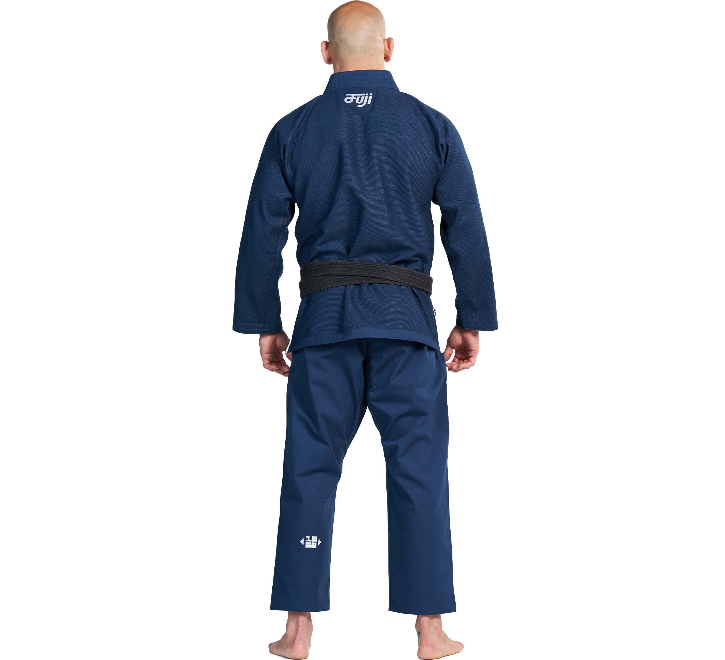 A person wearing the Flow-Tech BJJ Gi Navy with a black belt stands barefoot, facing away from the camera. The gi features white text on the upper back and lower right pant leg.