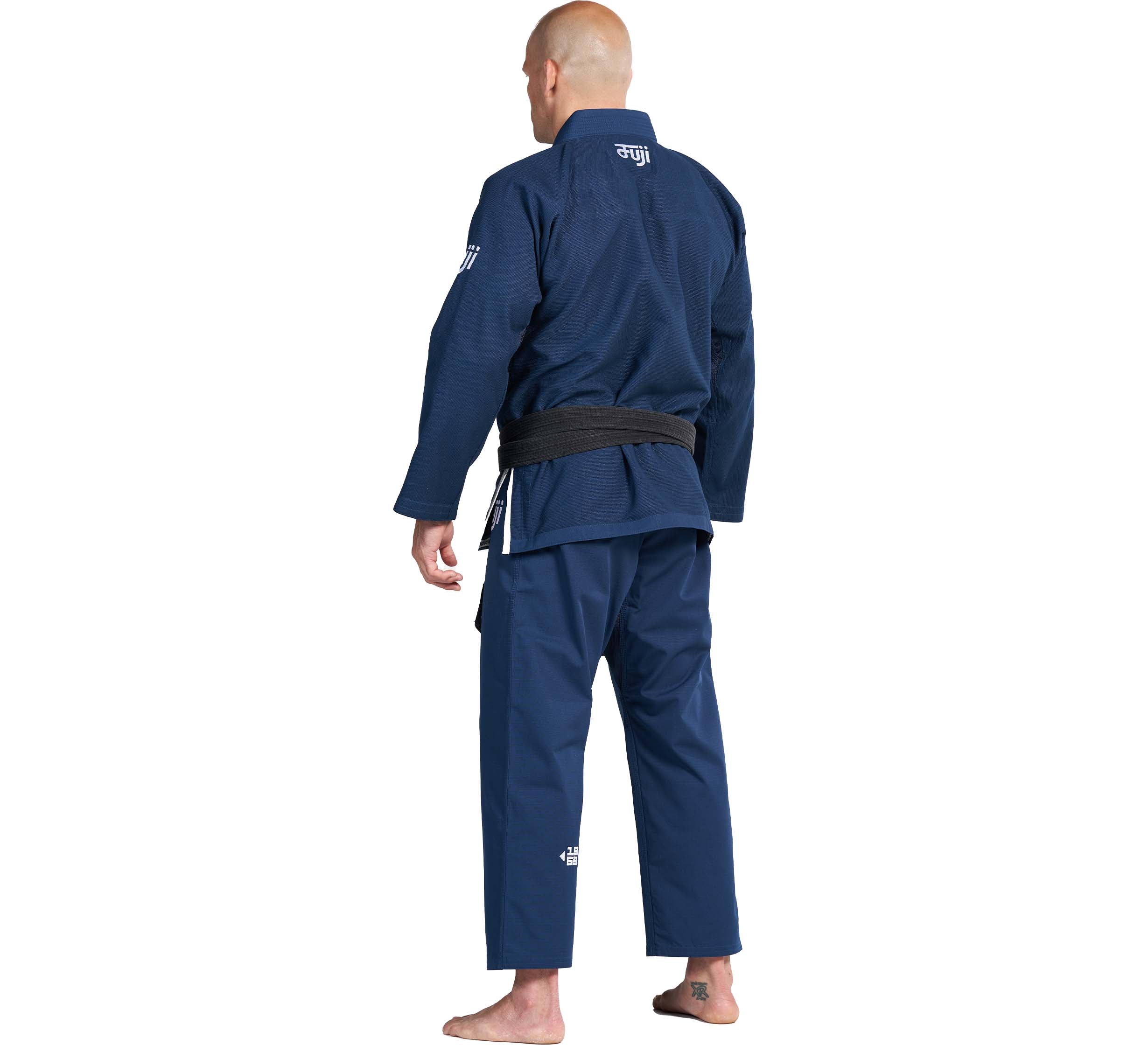 A person stands barefoot, facing away from the camera in a Flow-Tech BJJ Gi Navy with a black belt against a white background.