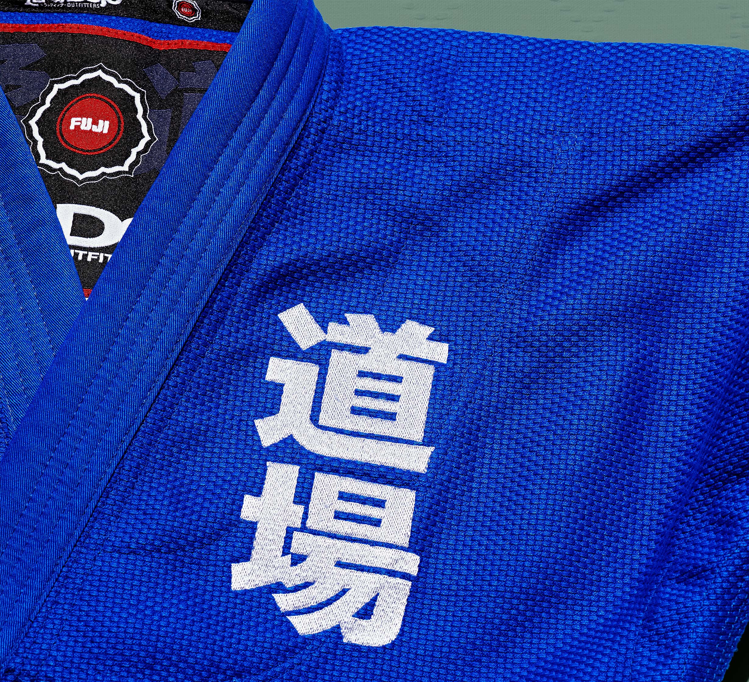 A close-up of the Dojo Outfitters x FUJI Judo Gi Blue shows white Japanese kanji embroidered on the chest, a FUJI logo on the collar, and detailed textured fabric with prominent stitching.