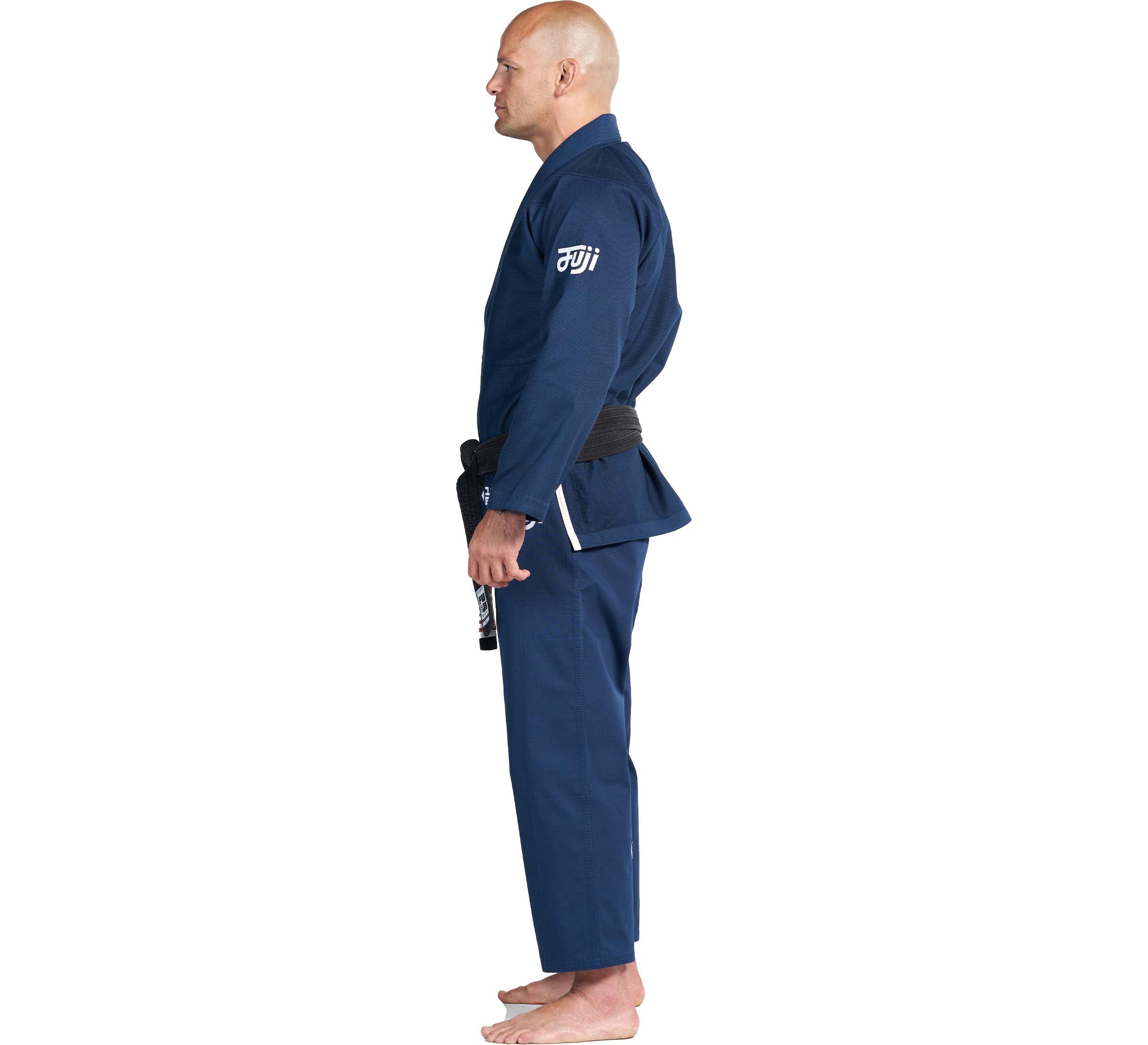 A man wearing the Flow-Tech BJJ Gi Navy with a black belt stands barefoot in profile against a white background.