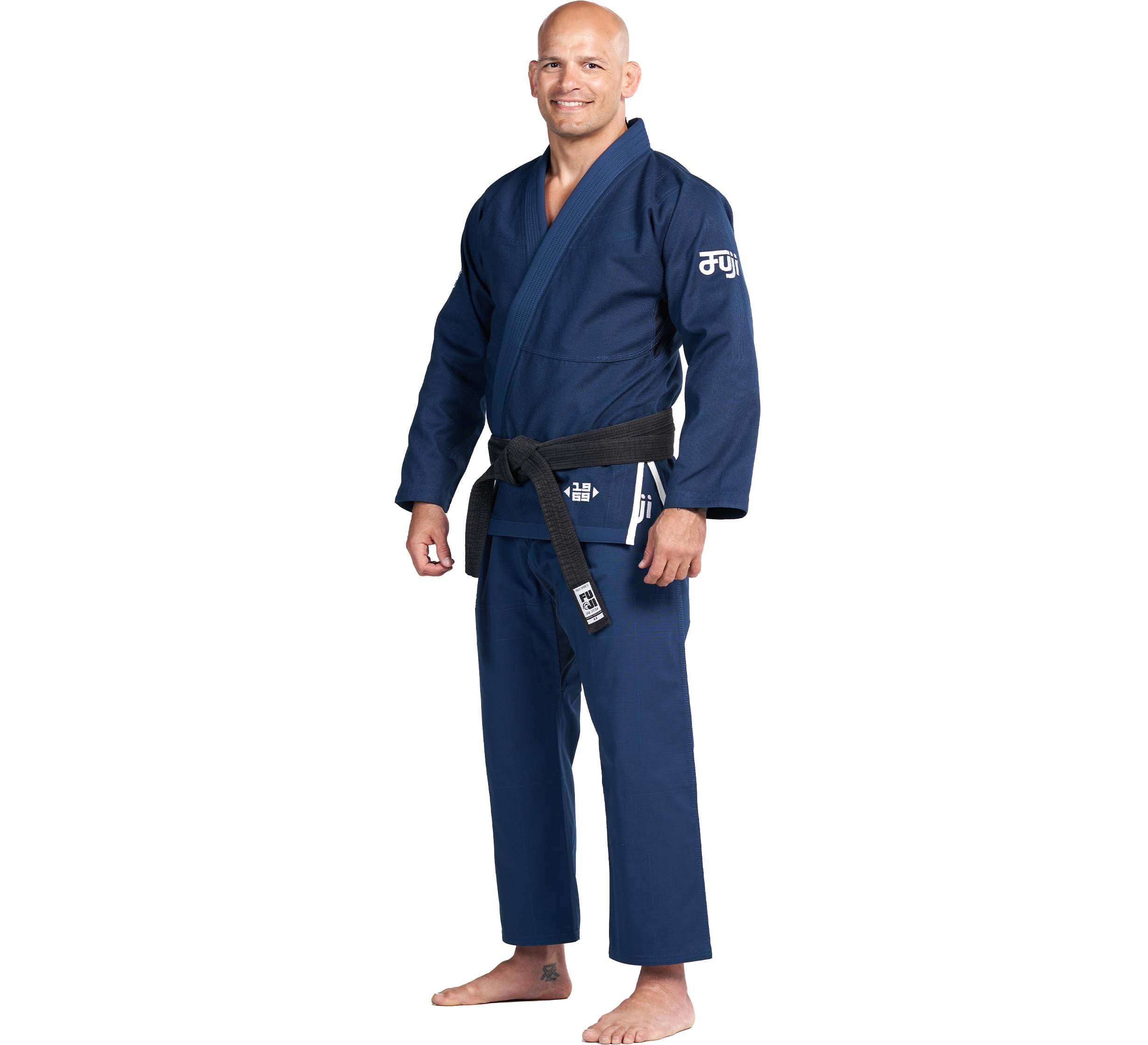 A man smiles barefoot in a Flow-Tech BJJ Gi Navy with a black belt, standing and facing slightly to the side against a plain white background.