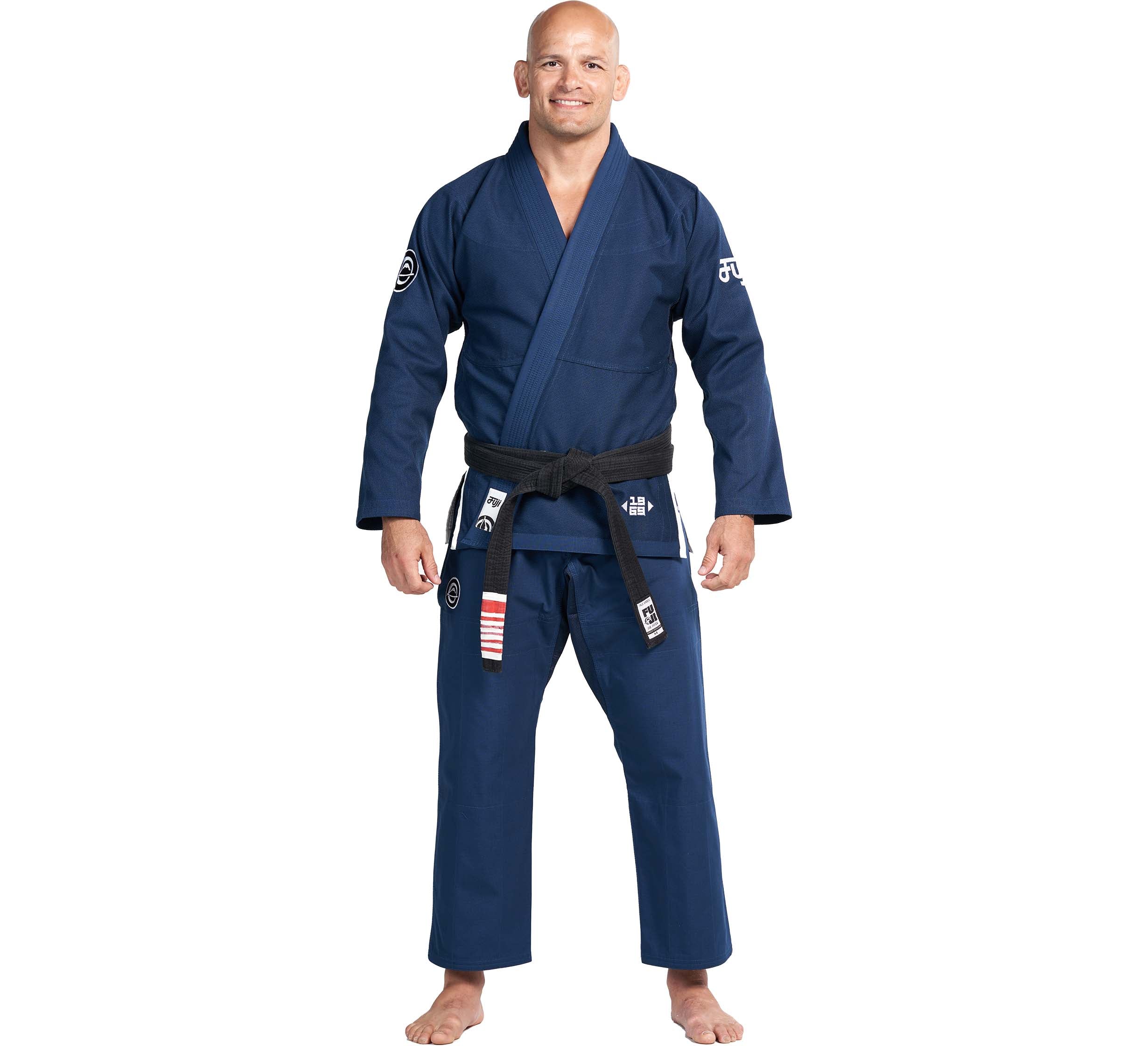 A bald man smiles barefoot at the camera, wearing the Flow-Tech BJJ Gi in navy with a black belt, standing against a plain white background.
