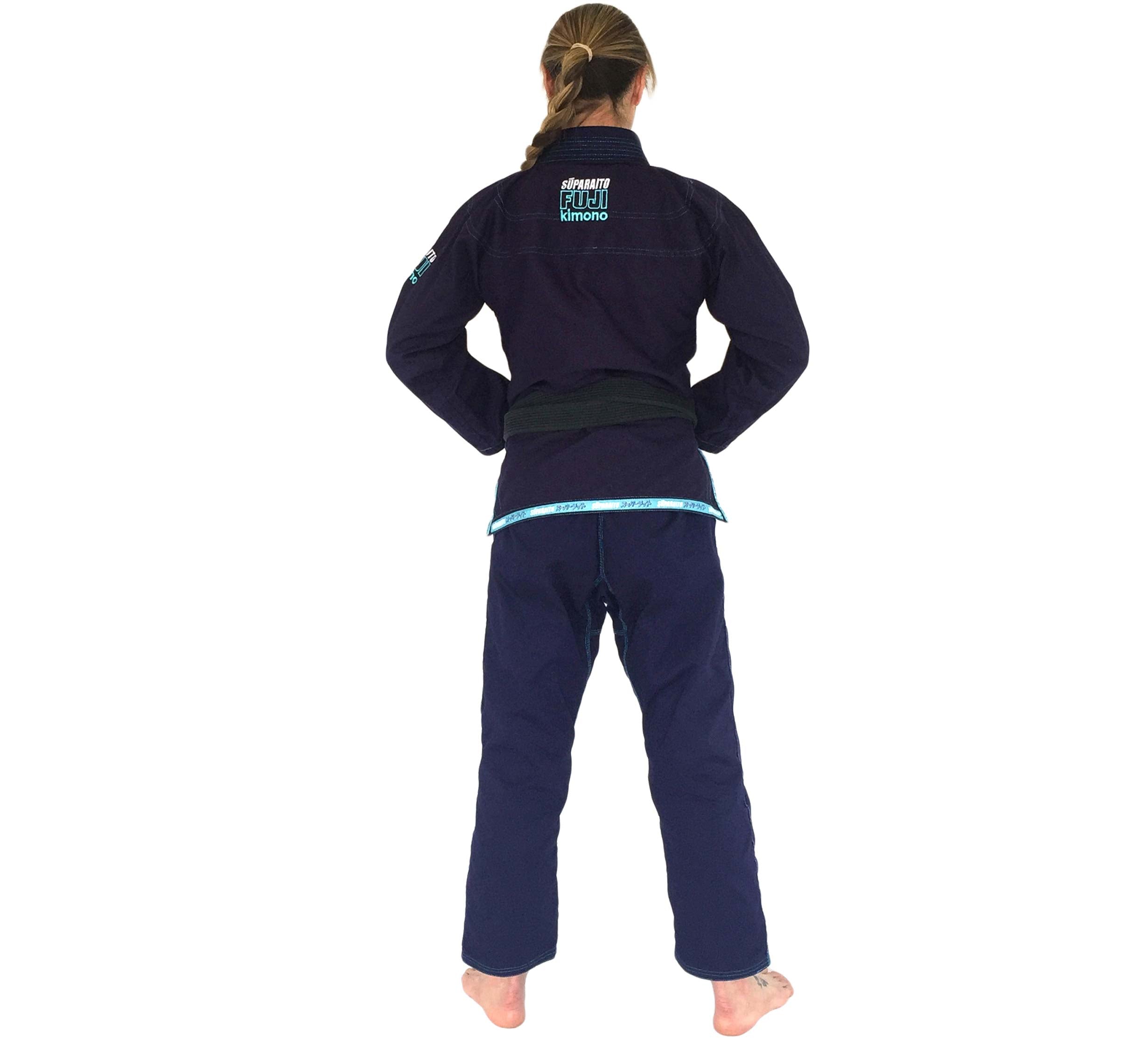 A barefoot woman with long hair faces away, wearing the Suparaito Womens BJJ Gi in navy with a black belt, set against a white background.
