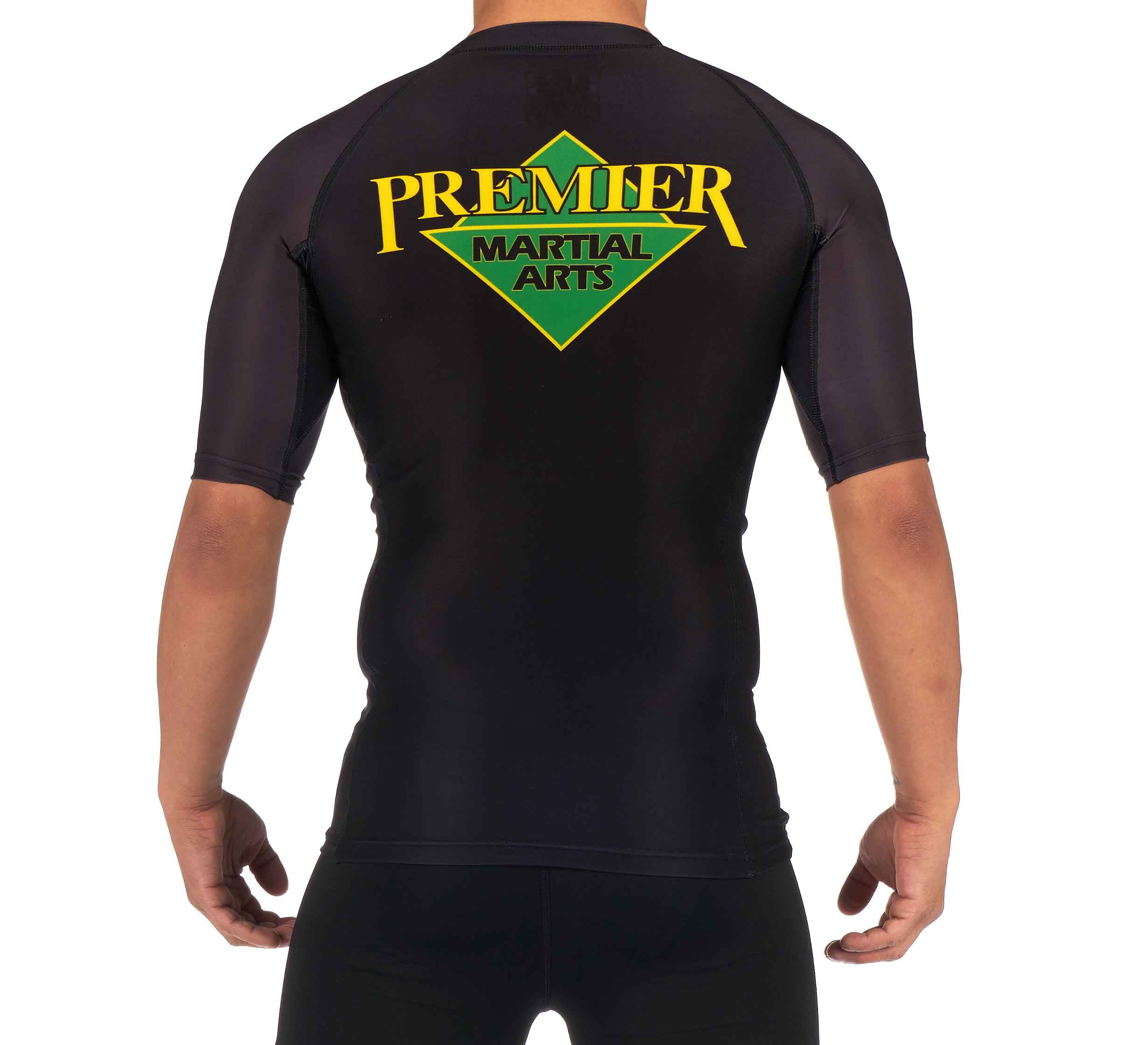 A child is seen from the back wearing the Premier Martial Arts Short Sleeve Kids Rashguard in black, featuring "Premier Martial Arts" in yellow and green on the back.