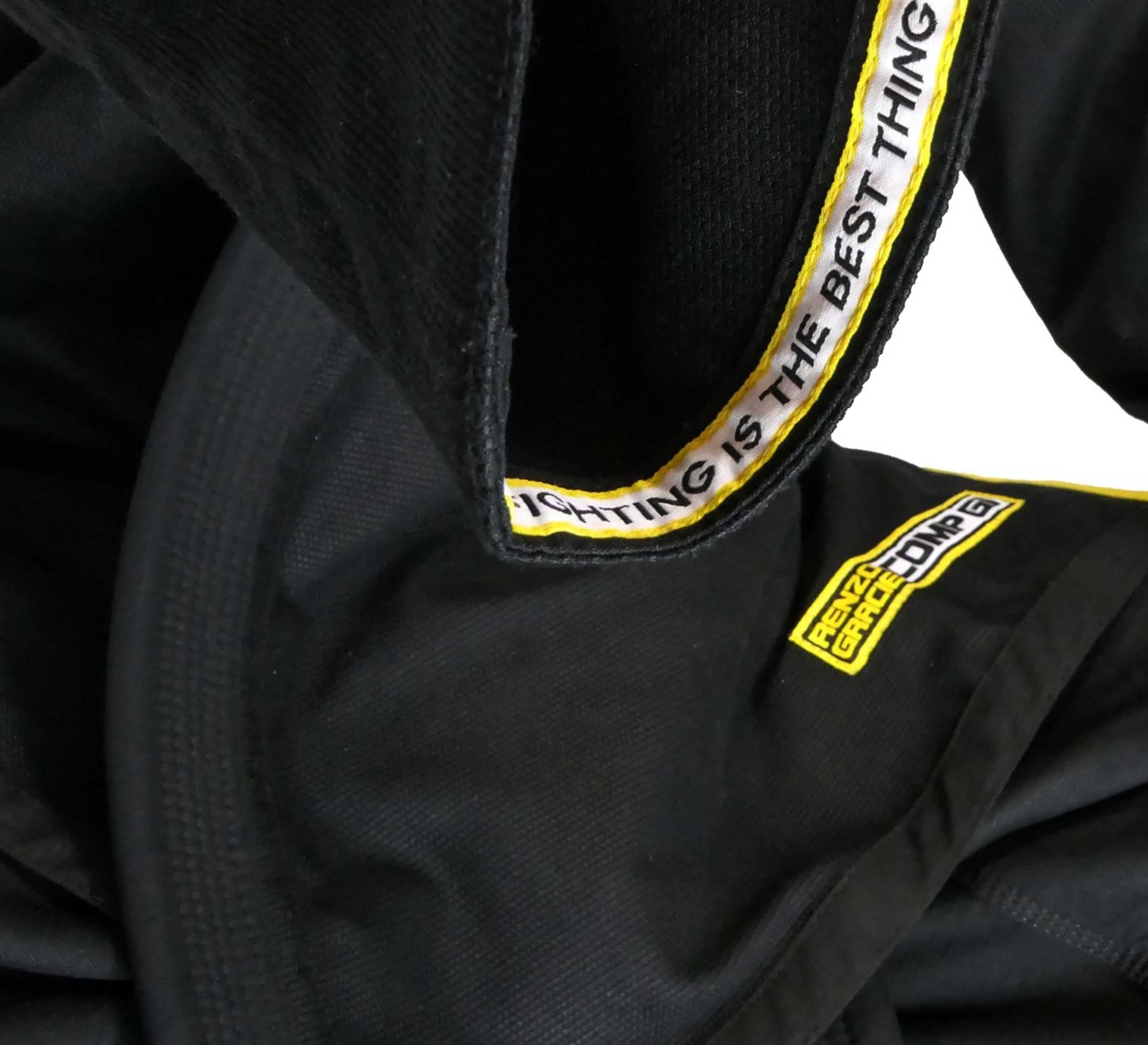 Close-up of the Renzo Gracie Superlite Gi, a black martial arts uniform with yellow trim. The collar features “FIGHTING IS THE BEST THING” text, a RIVAL GEAR CO. UNG patch, and detailed fabric texture.