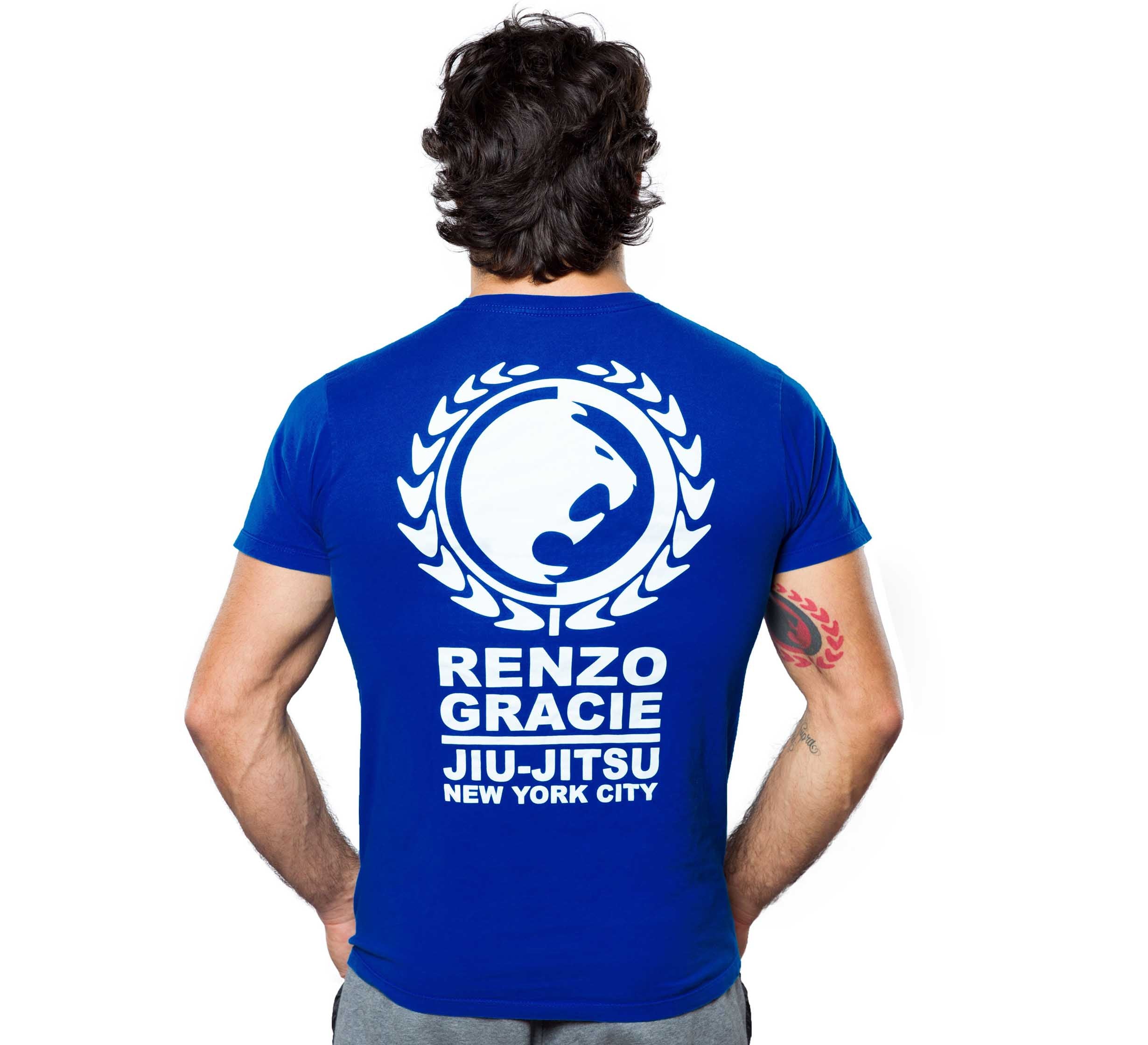 A man with dark, wavy hair faces away, wearing the Renzo Gracie Classic Ranked T-shirt in blue. The back features a white New York City logo with a laurel design. He has a red tattoo on his right arm.