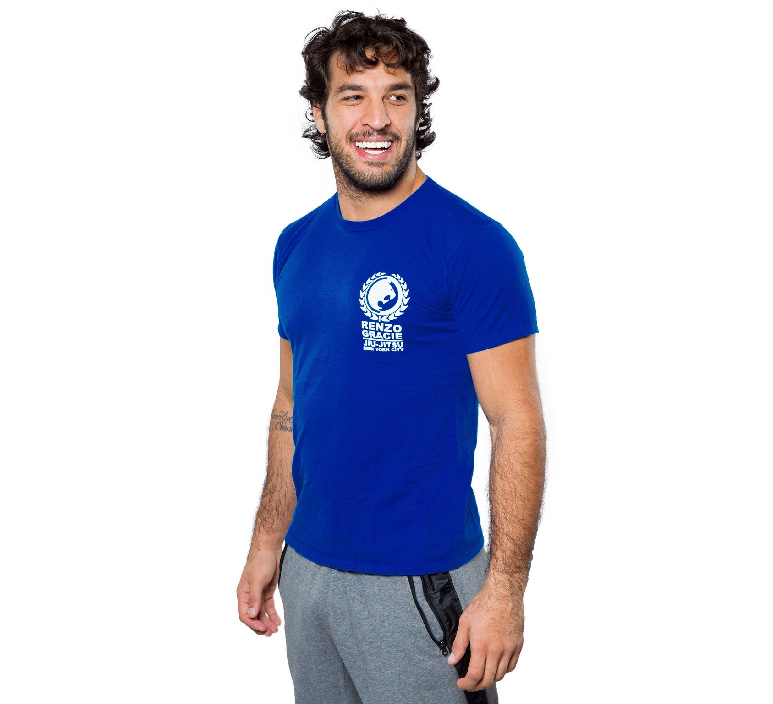 A smiling man with curly hair wears a blue Renzo Gracie Classic Ranked T-shirt and gray sweatpants, standing against a plain white background.