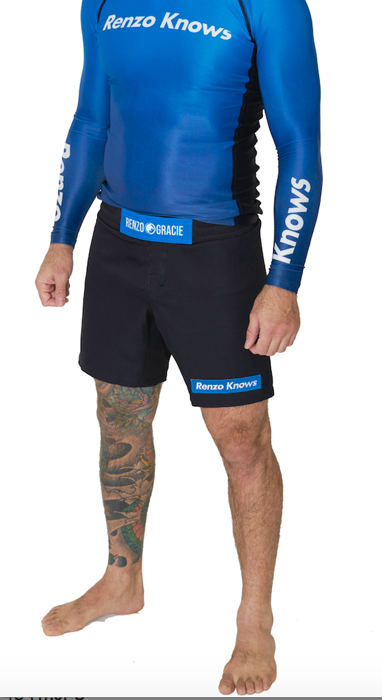A person stands barefoot against a white background wearing blue long-sleeve rash guard and Renzo Gracie Limited Edition Renzo Knows Fight Shorts Blue/Black, with one leg tattooed and visible Renzo Knows and Renzo Gracie logos.