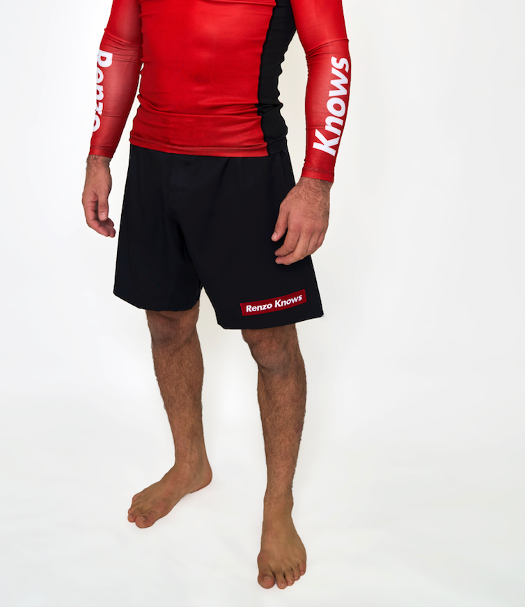 A barefoot man wearing a red and black long-sleeve rash guard and Renzo Gracie Limited Edition Renzo Knows Fight Shorts Black/Red, standing against a plain white background.