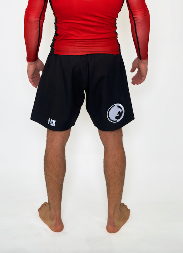 A person stands barefoot, facing away, wearing a red long-sleeve top and Renzo Gracie Limited Edition Renzo Knows Fight Shorts in black/red with white logos on the back against a plain white background.
