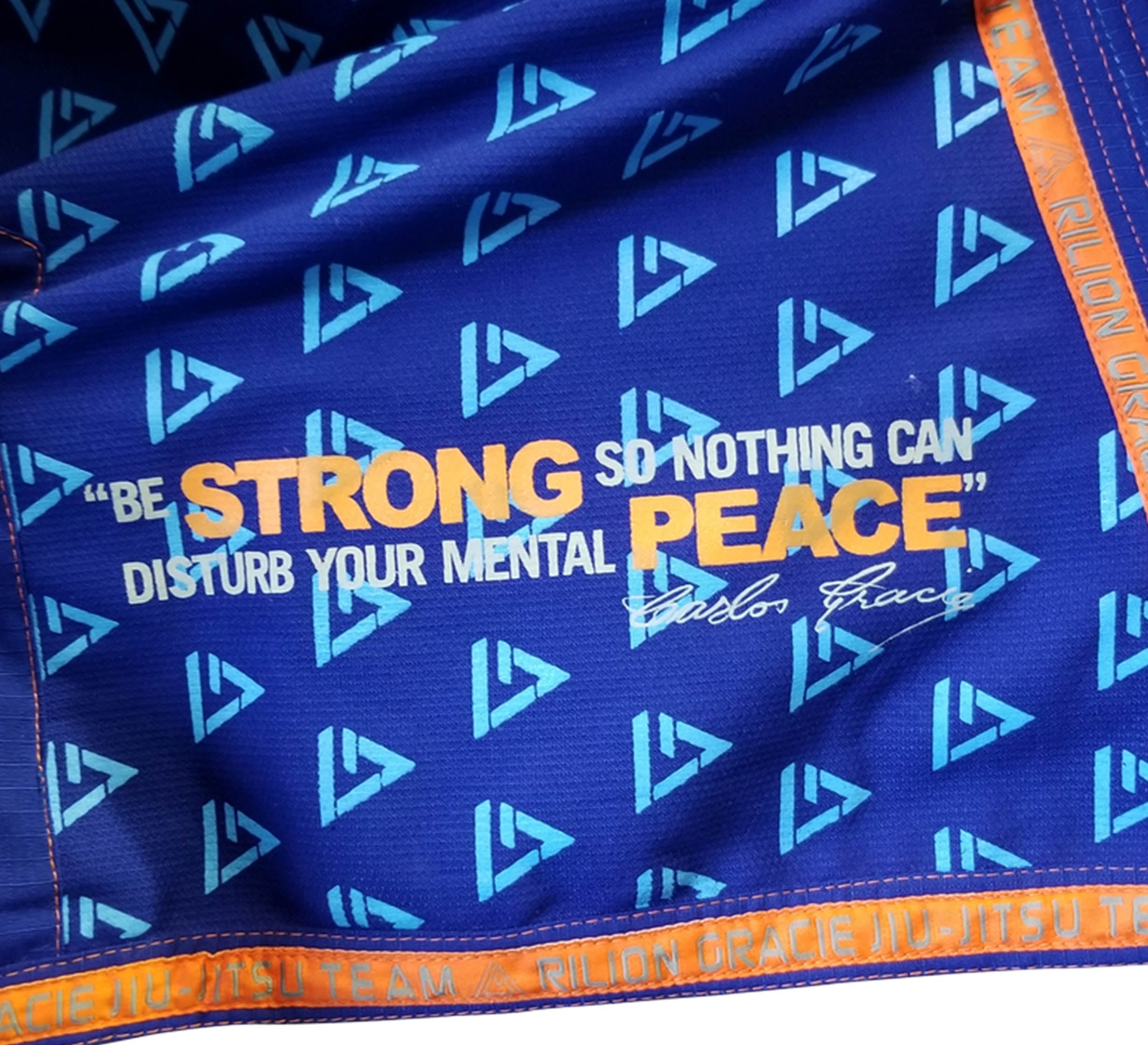 The Rilion Gracie Superlite Gi features blue fabric with repeating white triangles, a quote by Carlos Gracie in white and yellow—"Be strong so nothing can disturb your mental peace"—and is finished with orange trim.