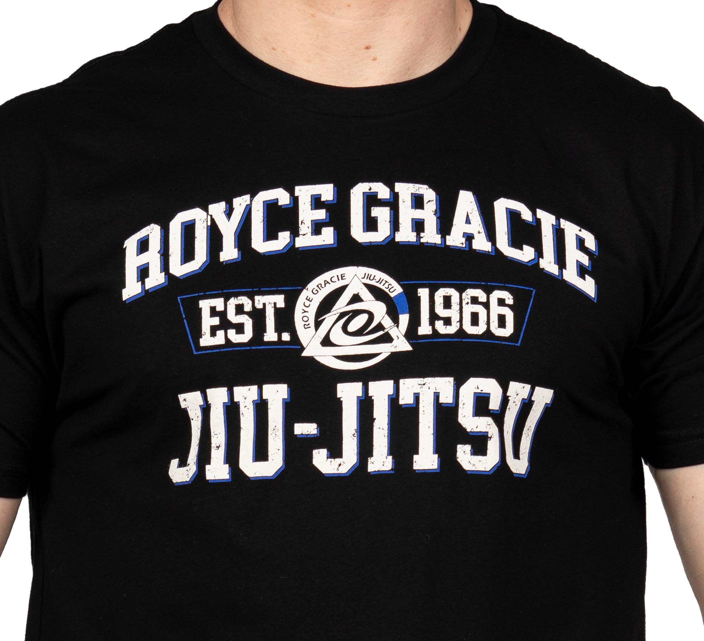 A person wearing the Royce Gracie Collegiate T-Shirt, which is black with "Royce Gracie Est. 1966 Jiu-Jitsu" in white and blue lettering and features a circular logo in the center.