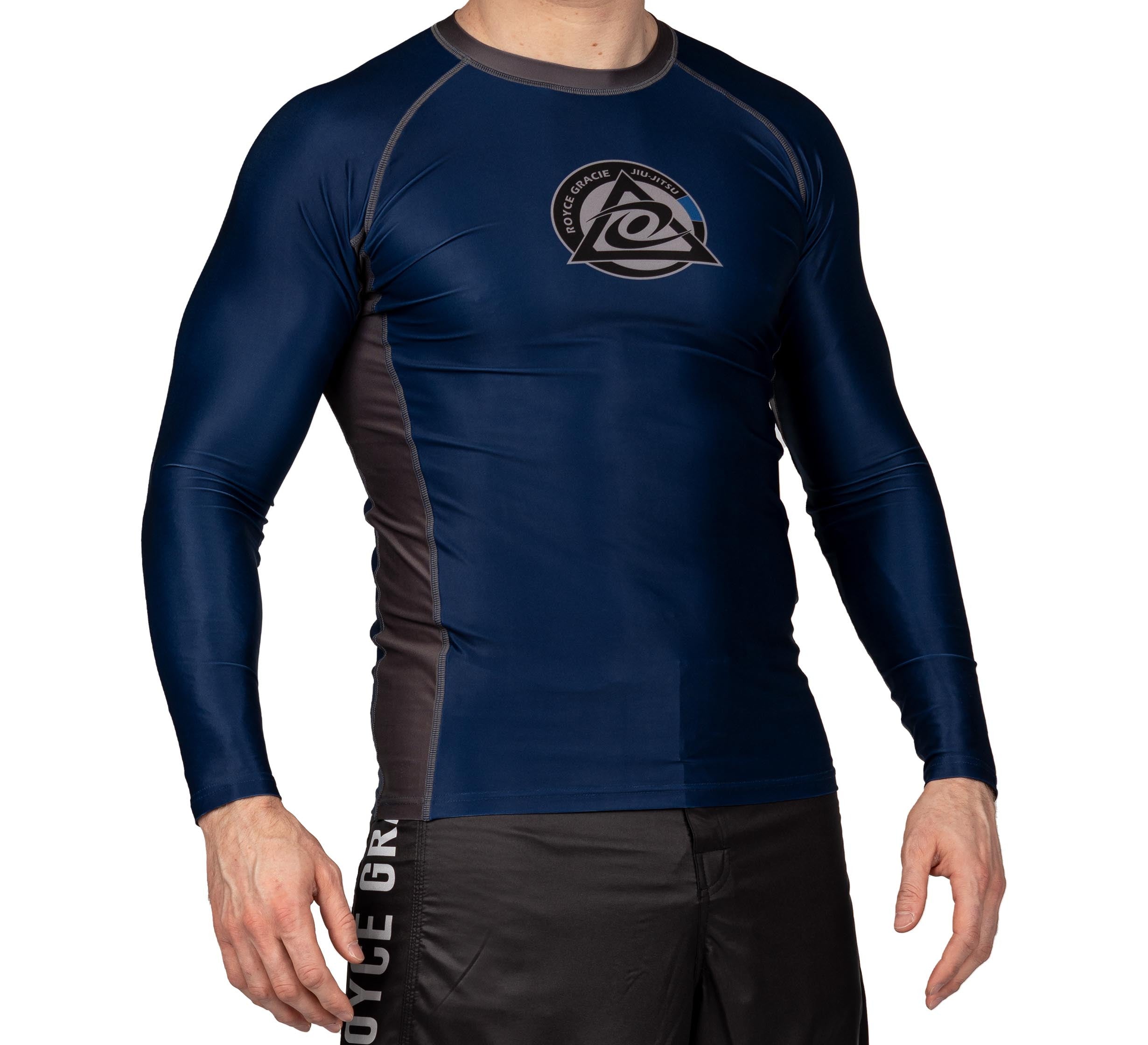A man wearing the Royce Gracie Official Long Sleeve Rashguard in navy and black, featuring a chest logo, paired with black shorts, stands against a white background. His head is not visible.