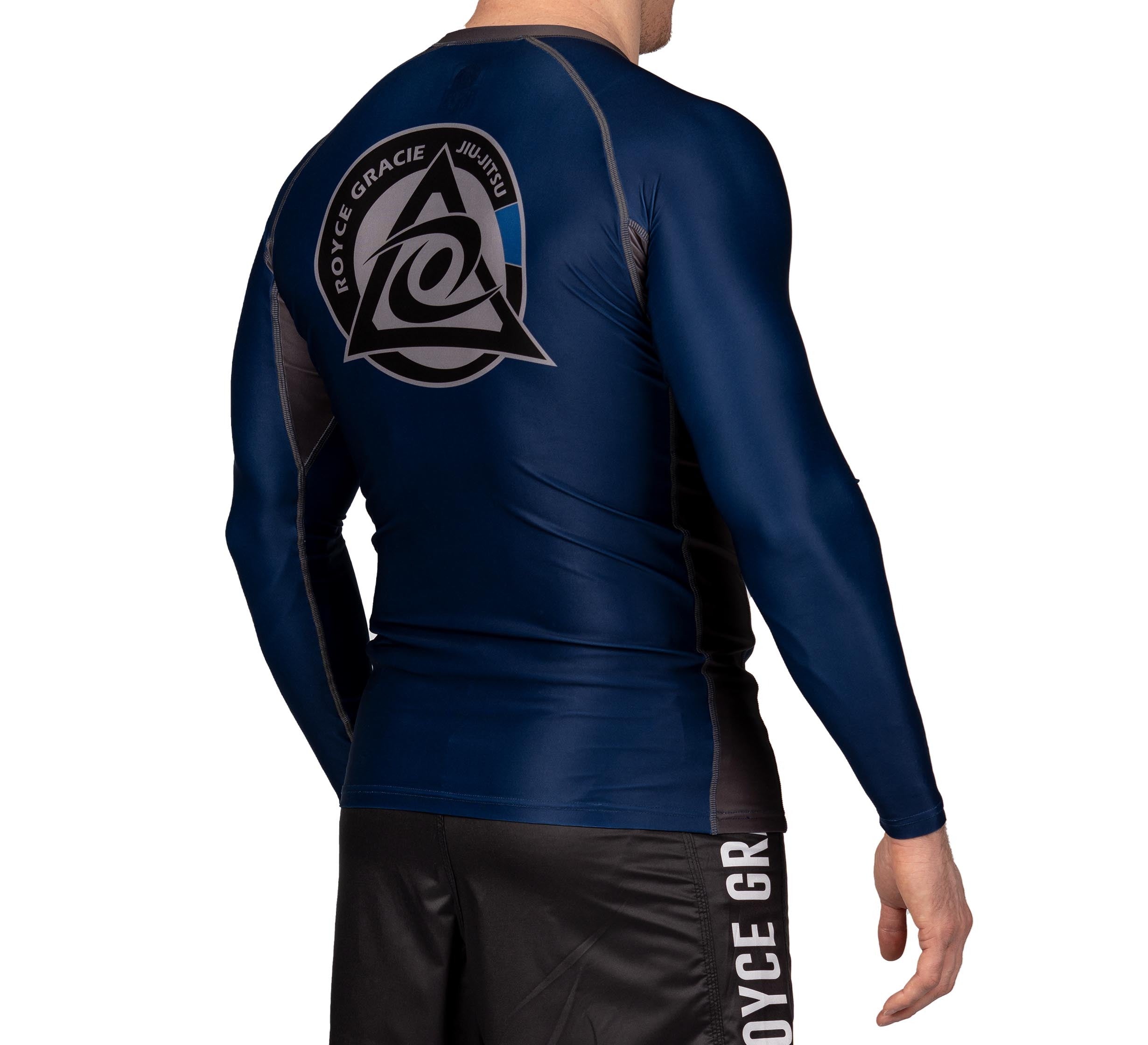 A child models the Royce Gracie Official Kids Long Sleeve Rashguard in dark blue, featuring the Royce Gracie Jiu-Jitsu logo on the back, paired with matching black shorts printed with “Royce Gracie,” against a white background.