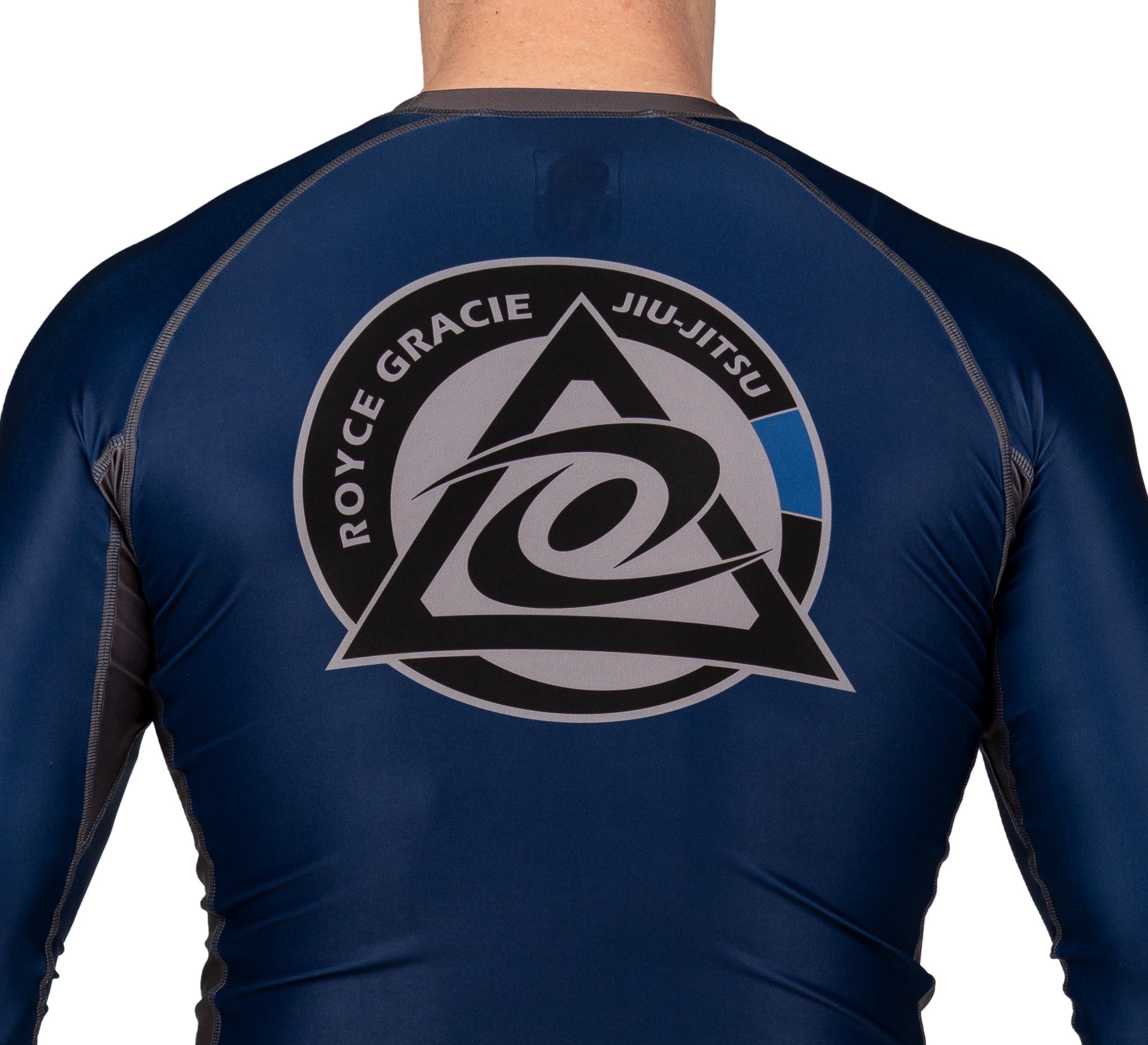 A child wears the Royce Gracie Official Kids Long Sleeve Rashguard in navy blue/black, featuring the Jiu-Jitsu logo with a stylized triangle and swirl on the back. The child's head is not visible.