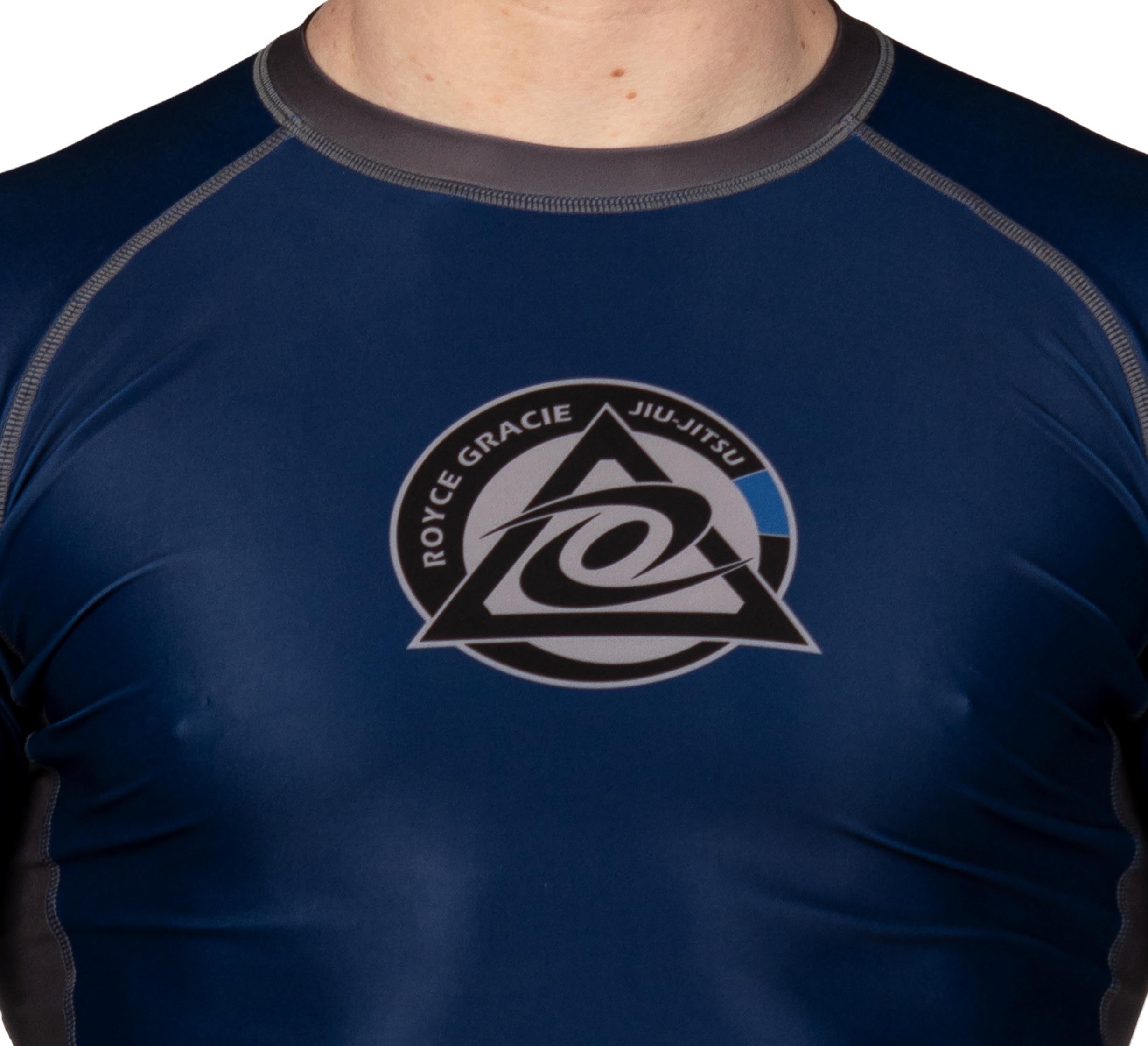 A child wears the Royce Gracie Official Kids Long Sleeve Rashguard in dark blue, featuring a triangle and stylized logo with circling text on the chest; only the upper torso and neck are visible.