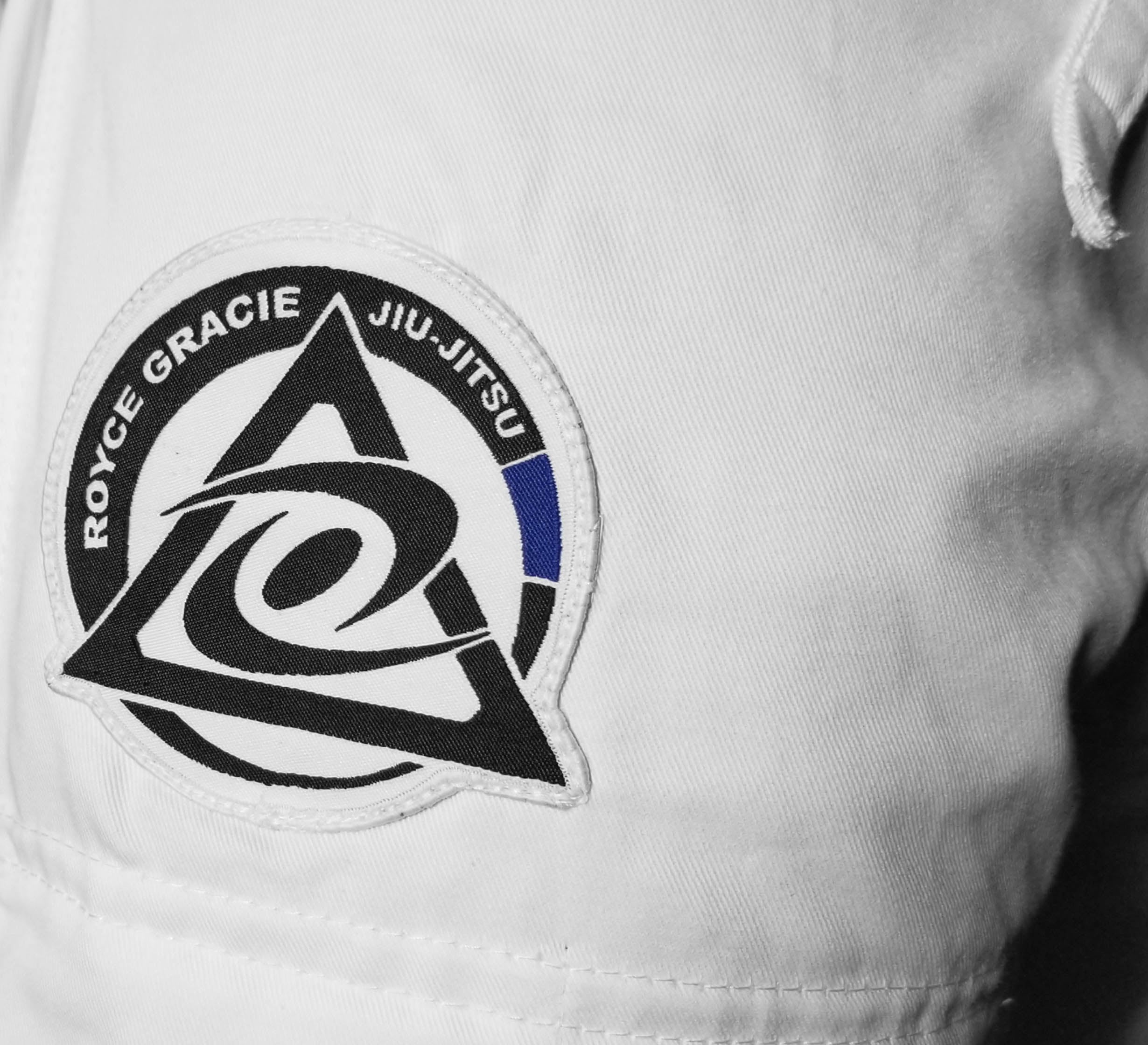 A close-up of the Official Royce Gracie Kids BJJ Gi White, featuring a patch with "Royce Gracie Jiu-Jitsu," a black triangle and swirl design, and a blue stripe on the right side.