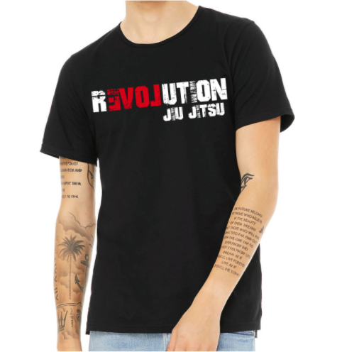 A person with visible arm tattoos stands against a plain background, wearing the BJJ Revolution Youth Black T-Shirt featuring "REVOLUTION JU JITSU" in bold white and red letters.