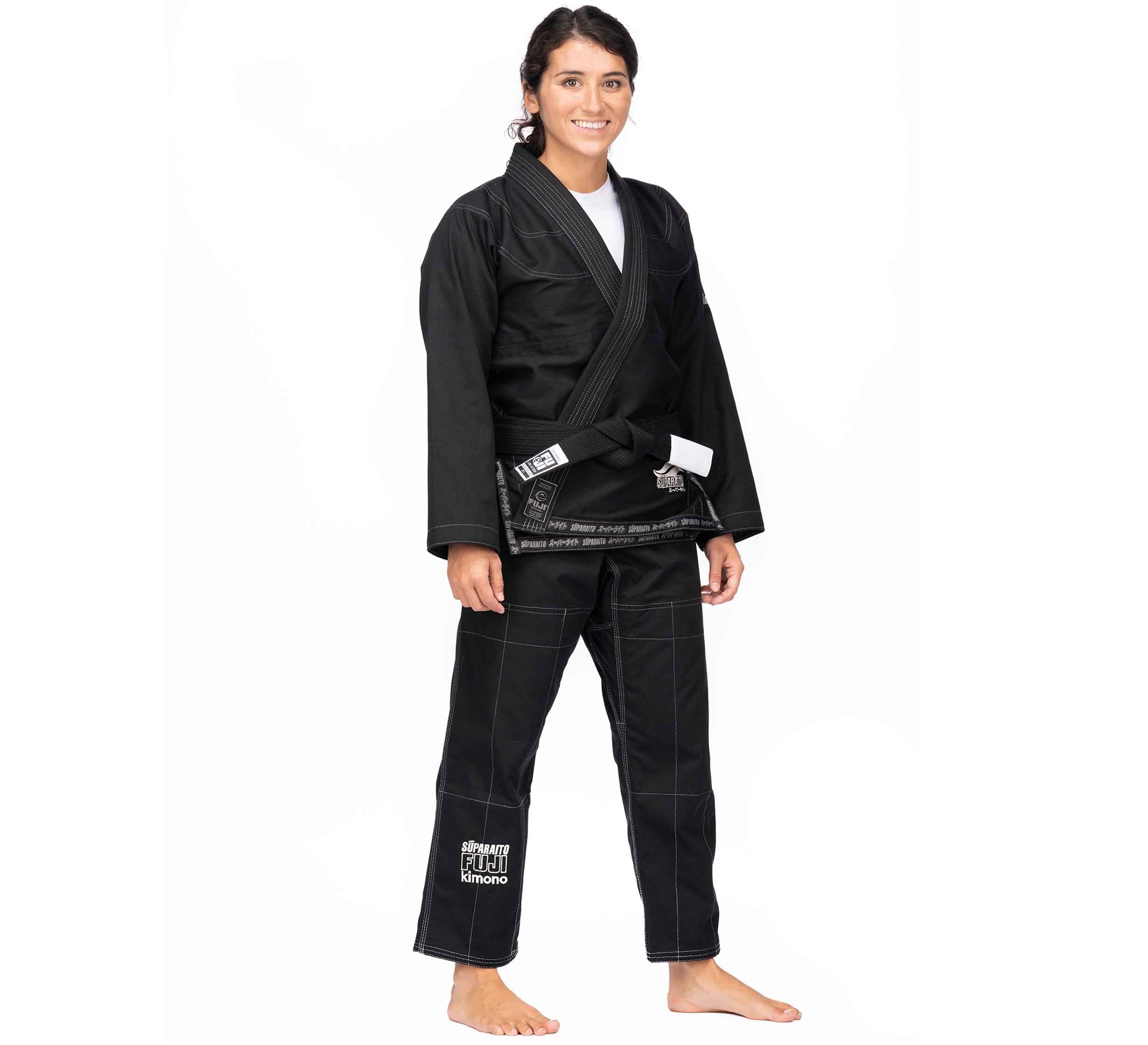 A person with long dark hair smiles barefoot in the Suparaito BJJ Gi (Unisex Size) Black and a black belt with white patch, standing against a plain white background.