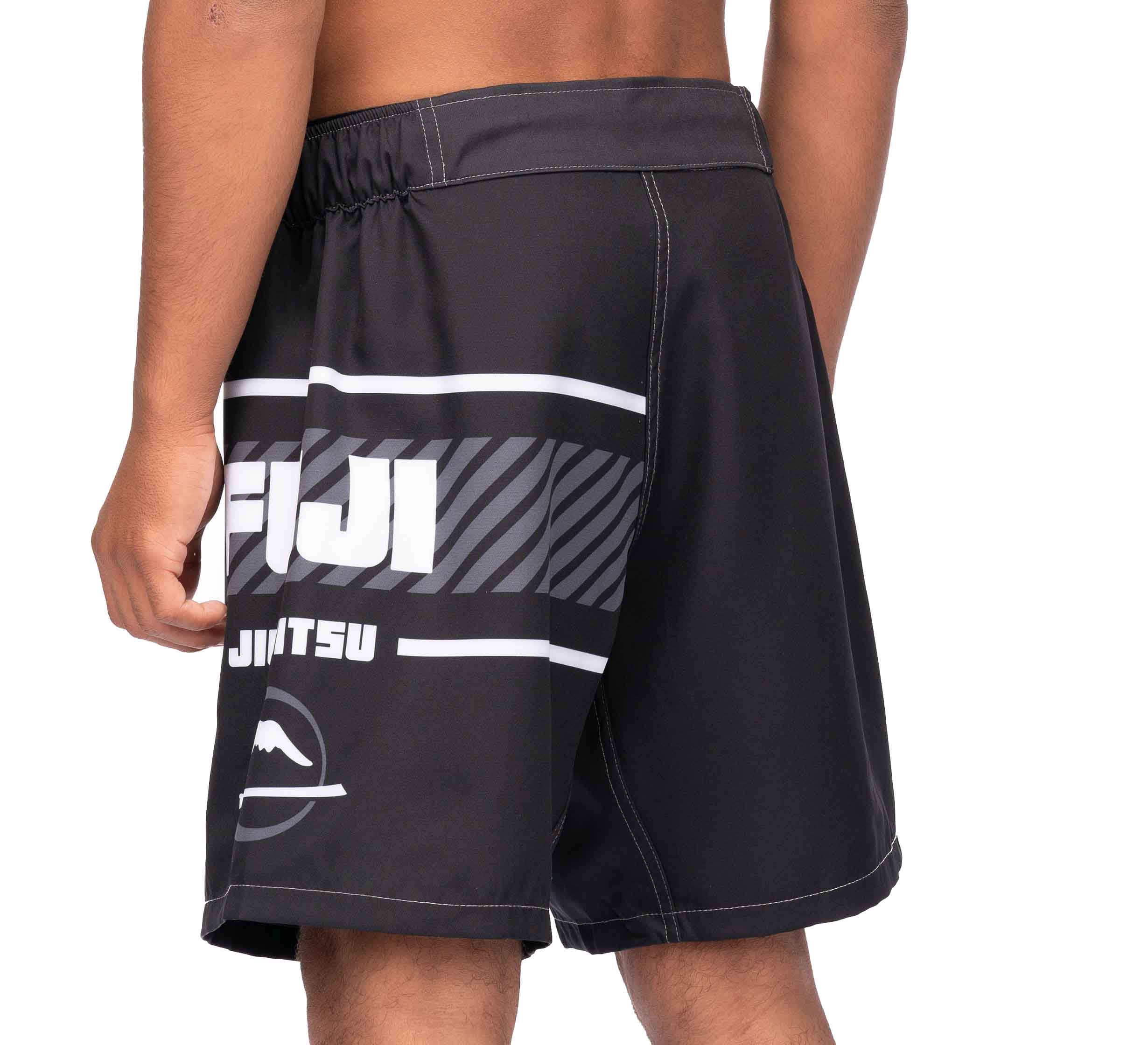 A person is standing sideways, showing the left side and back of the Freestyle 2.0 Ranked Grappling Shorts—black athletic shorts with white graphics and “FUJI” and “Jiu Jitsu” text on the side.