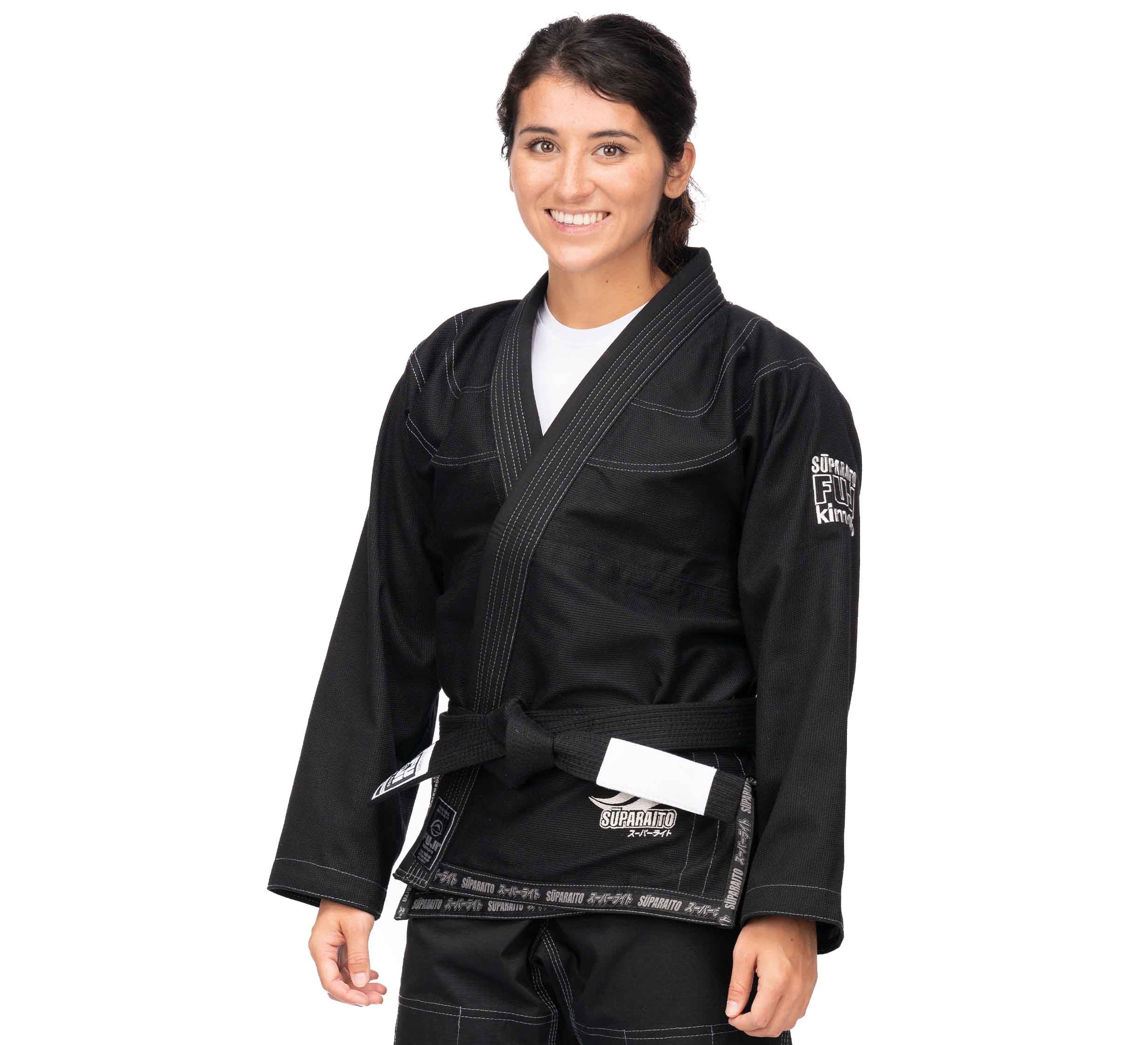 A woman smiles, wearing the Suparaito BJJ Gi (Unisex Size) in black with a black belt and white undershirt. The gi features patches on the sleeve and belt. Her dark hair is pulled back, and she stands against a white background.