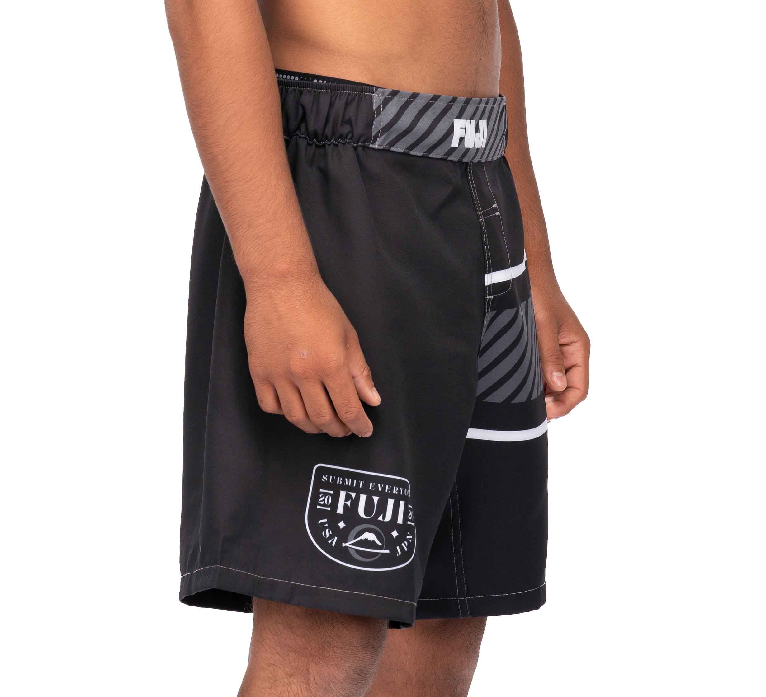 A person wearing black FUJI Freestyle 2.0 Ranked Grappling Shorts with white and gray accents, standing sideways with their right arm relaxed. The shorts display a logo and text near the hem.