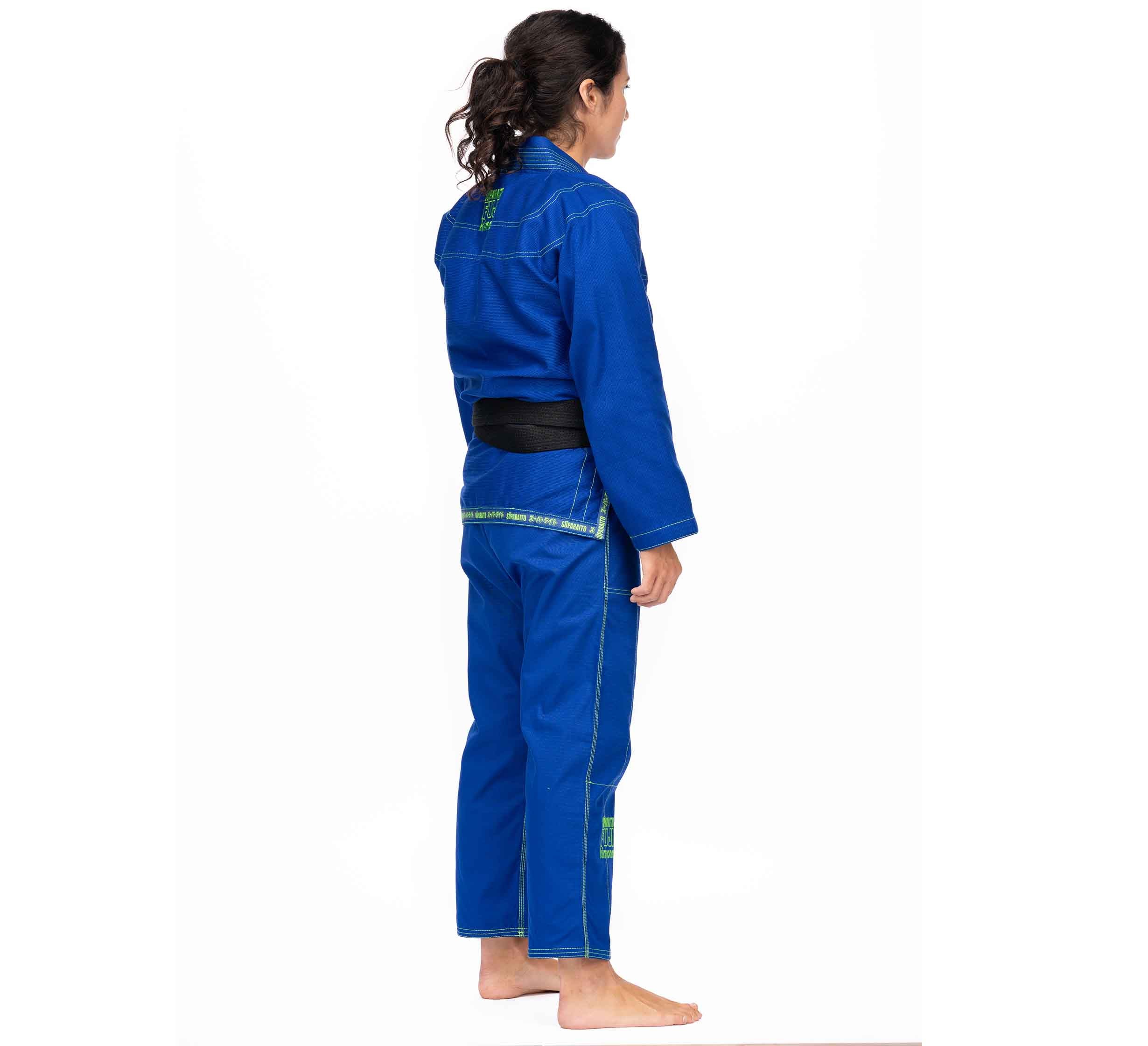 A person with long dark hair in a ponytail stands barefoot, wearing the Suparaito BJJ Gi (Unisex Size) in blue and a black belt, facing sideways with their left side visible against a plain white background.