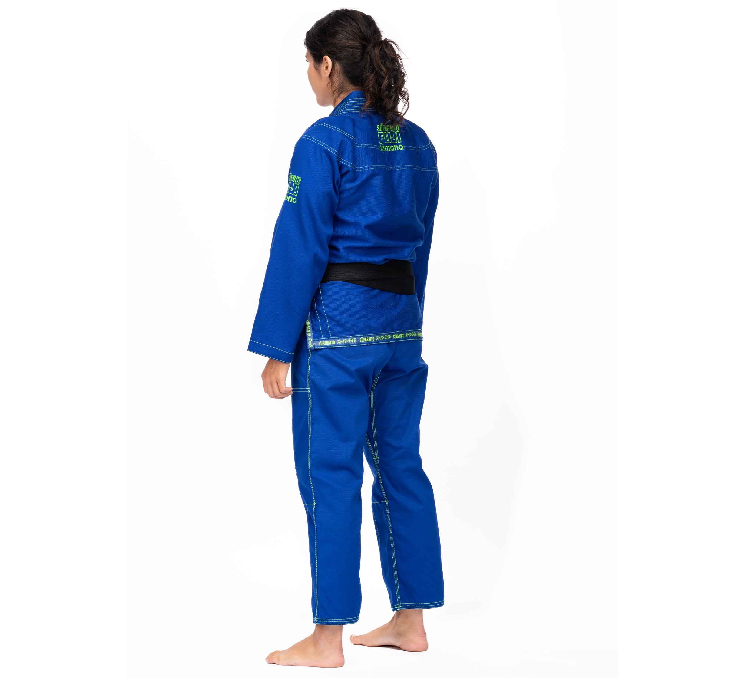 A barefoot person with long dark hair faces away, wearing a blue Suparaito BJJ Gi (Unisex Size) with green embroidered logos and a black belt, standing against a plain white background.