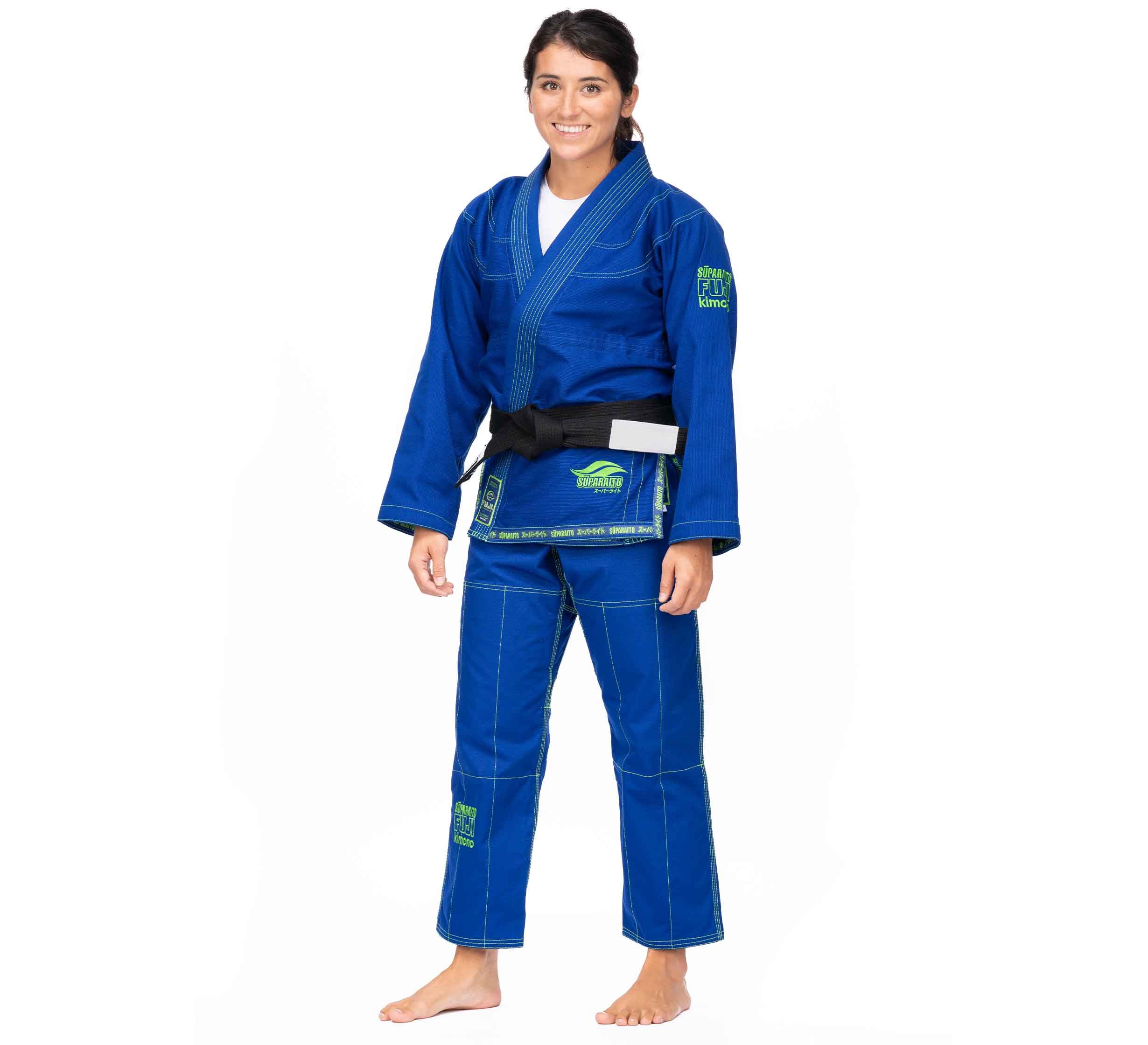 A woman stands barefoot, smiling in a Suparaito BJJ Gi (Unisex Size) Blue with green patches and a black belt against a plain white background.