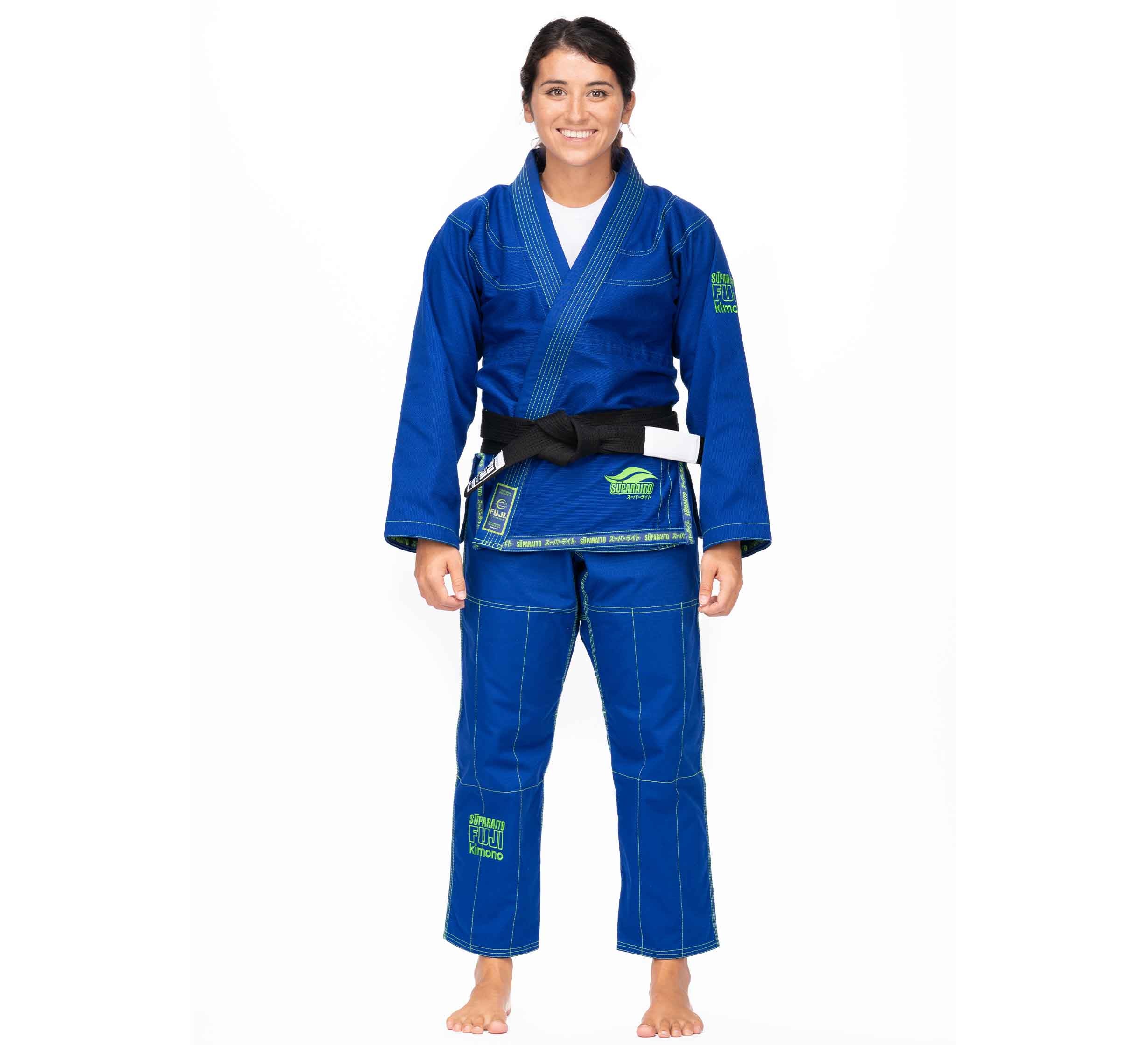 A woman stands barefoot, smiling, wearing the Suparaito BJJ Gi (Unisex Size) in blue with green patches and stitching, paired with a black belt. She poses against a plain white background.