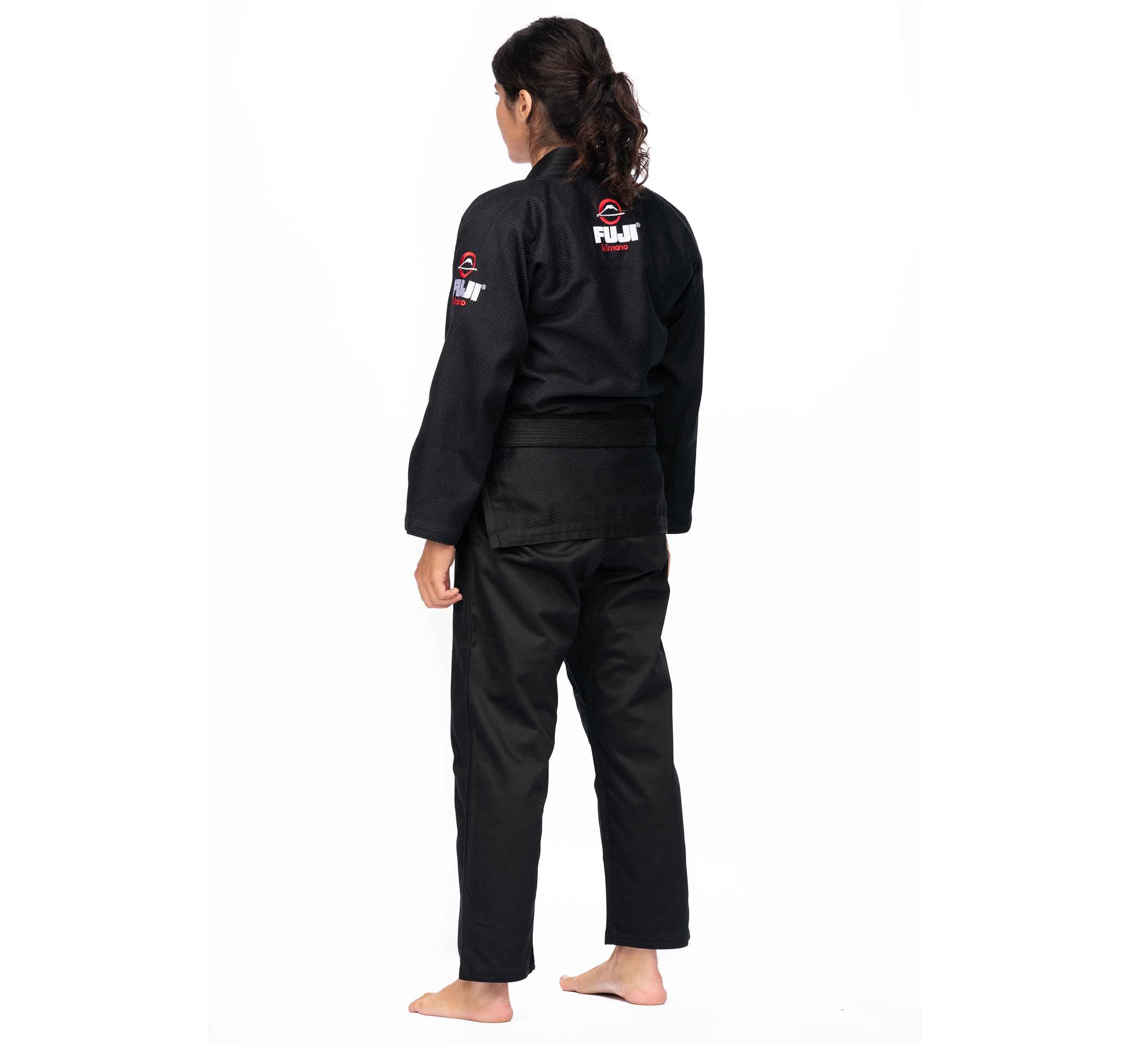 A person with long hair tied back stands barefoot, facing away, wearing the All Around BJJ Gi Black (Unisex Size) featuring FUJI logos on the upper arm and back, against a plain white background.