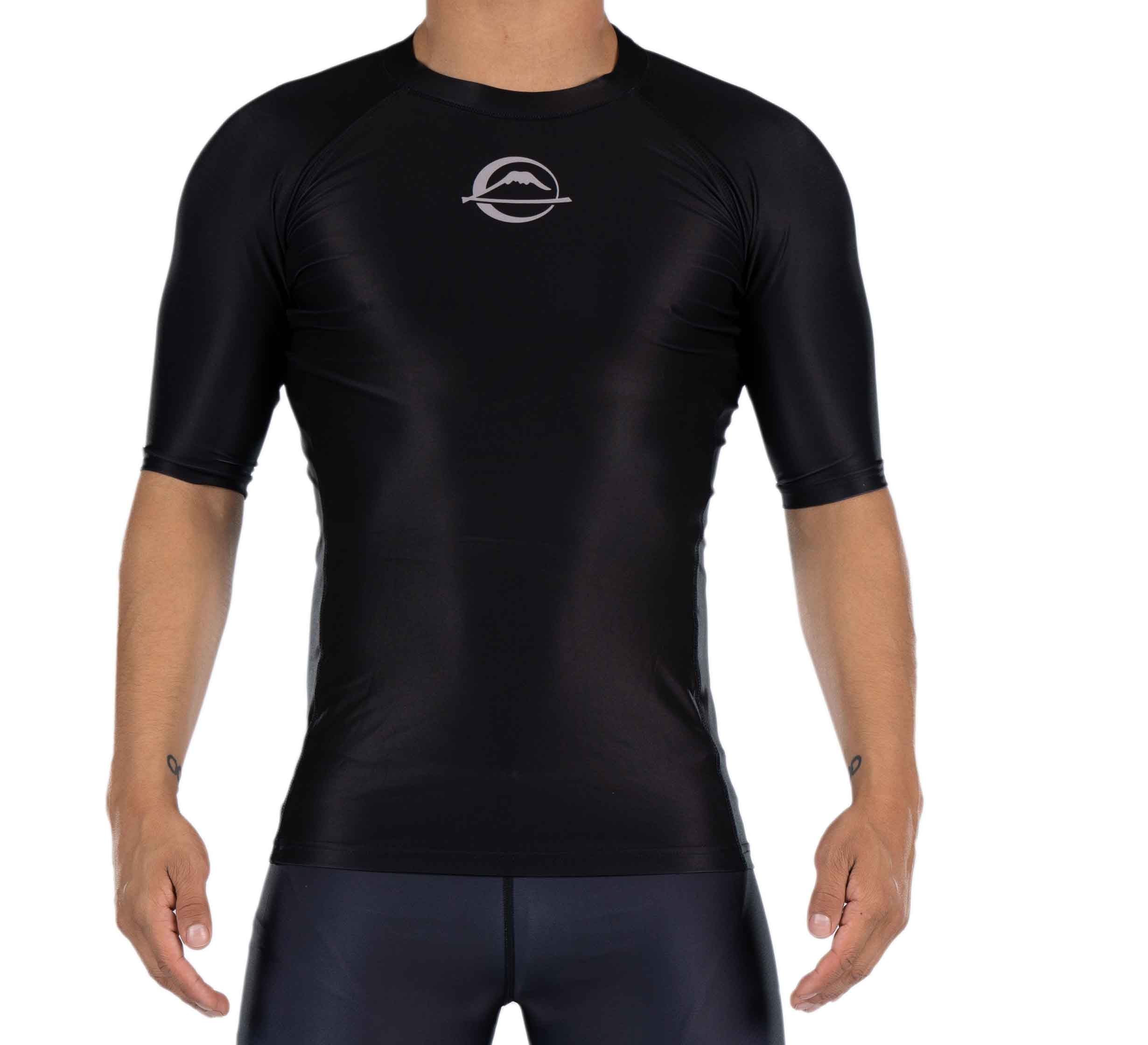 A person wearing the Baseline Ranked Short Sleeve Rashguard, a fitted black athletic shirt with a round chest logo and smooth, shiny finish, stands with arms slightly out to the sides.
