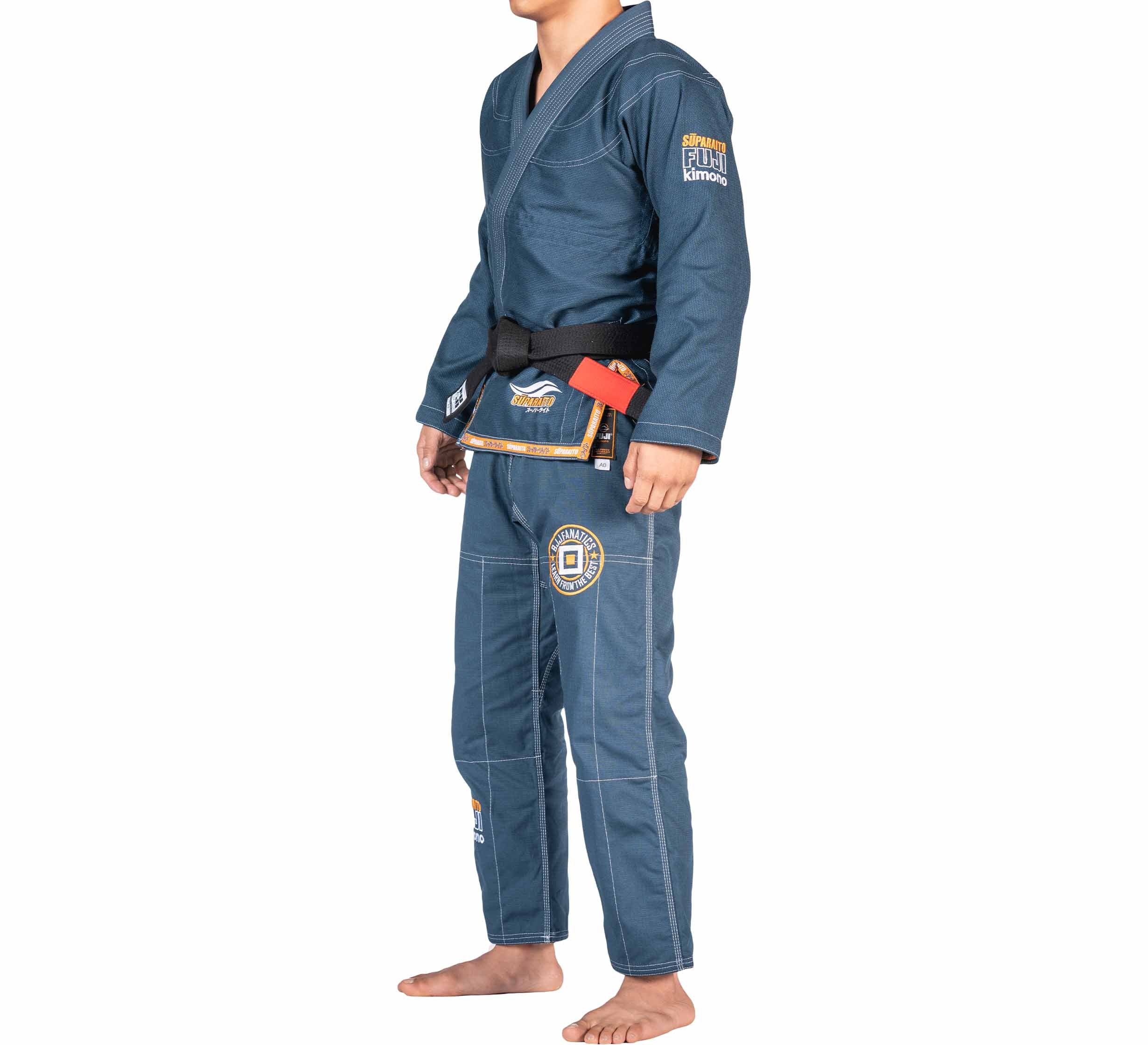 A person stands barefoot in the FUJI X BJJ Fanatics Blue Suparaito Gi with patches and a black belt, facing slightly to the side against a white background.