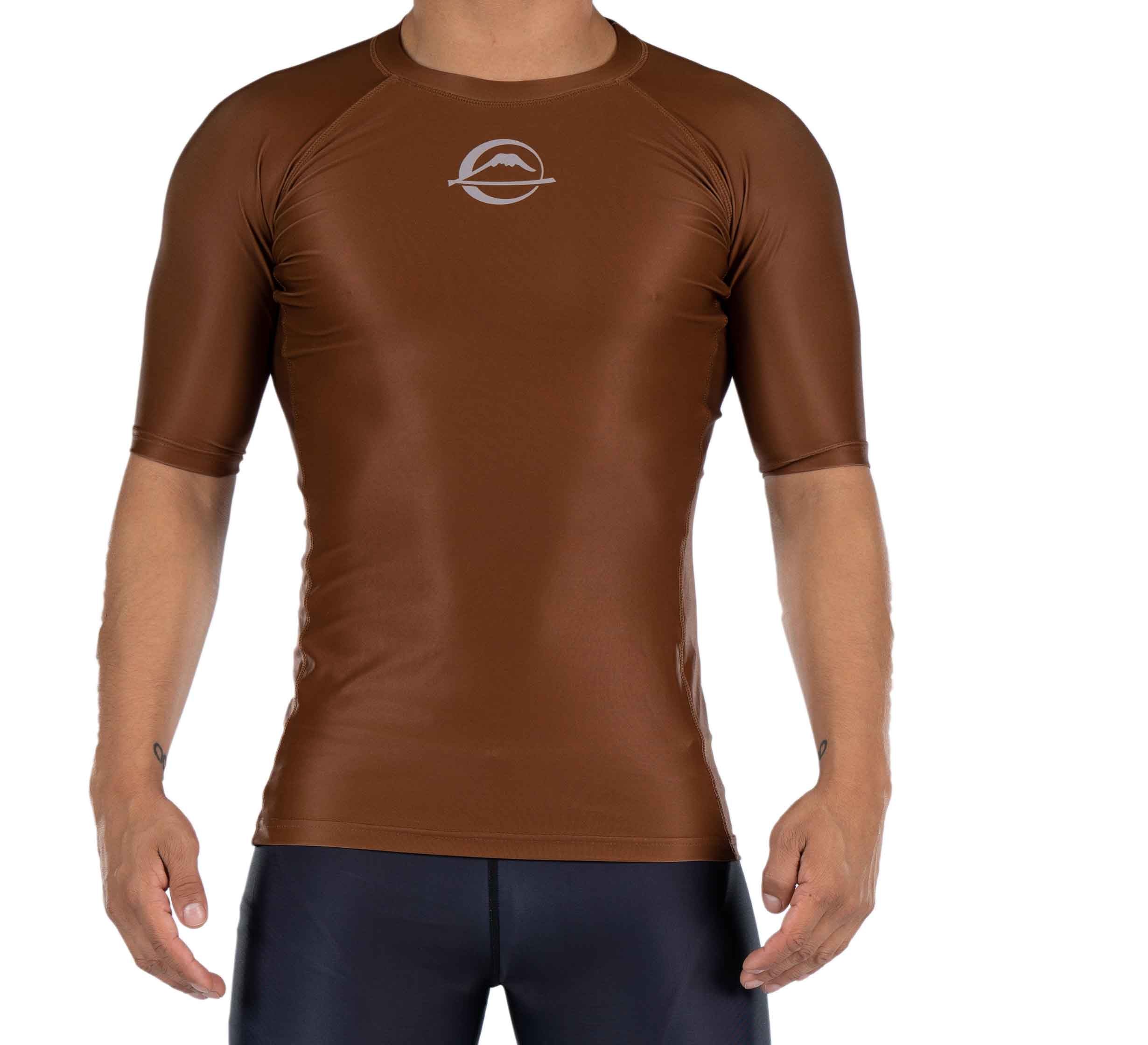 A person wears the Baseline Ranked Short Sleeve Rashguard Brown, a fitted, short-sleeved athletic shirt with a small white chest logo, paired with dark shorts and standing against a white background.
