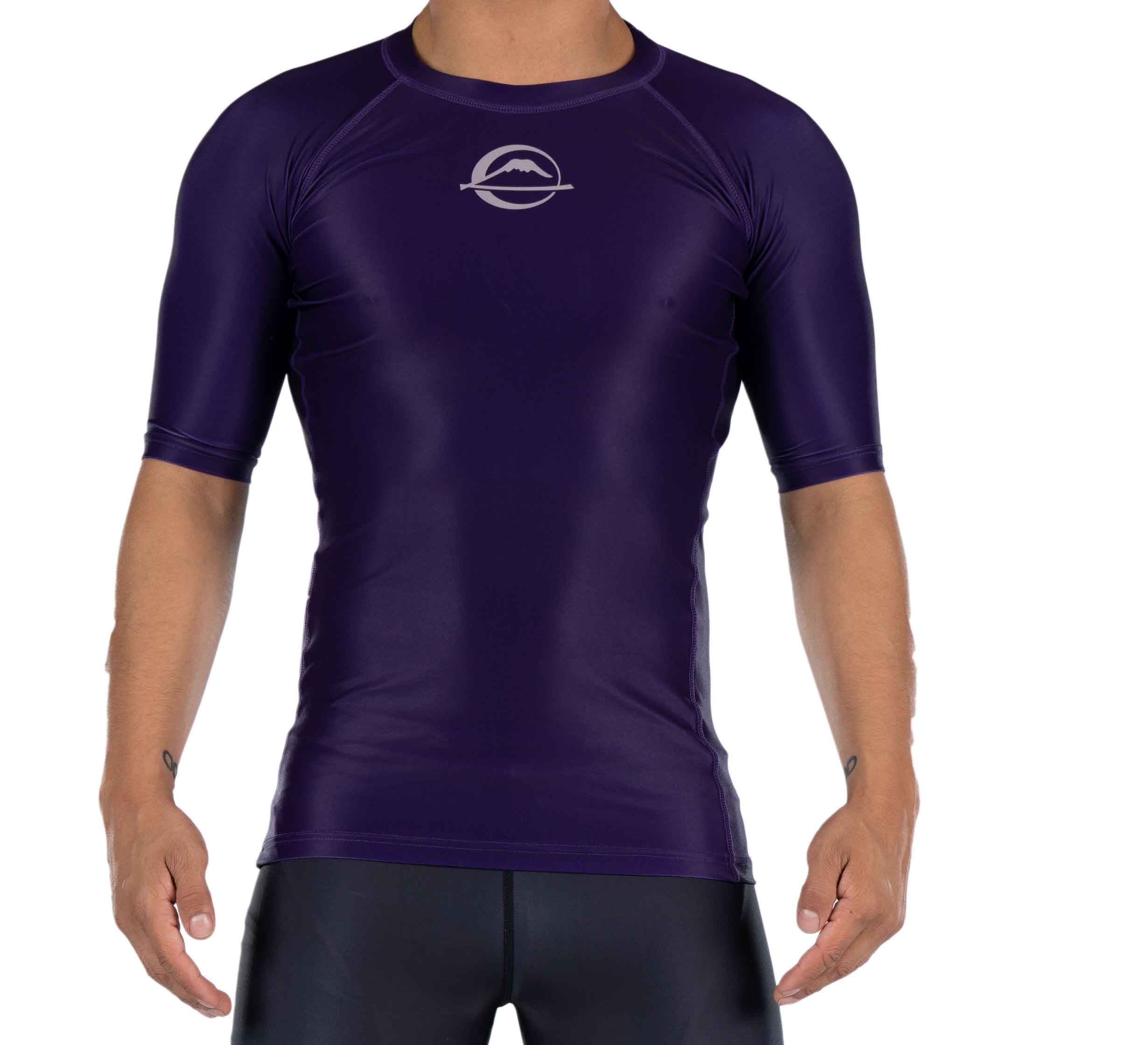 A person wearing the Baseline Ranked Short Sleeve Rashguard in purple, featuring a tight fit, short sleeves, and a white circular mountain logo on the chest, stands with arms relaxed by their sides.