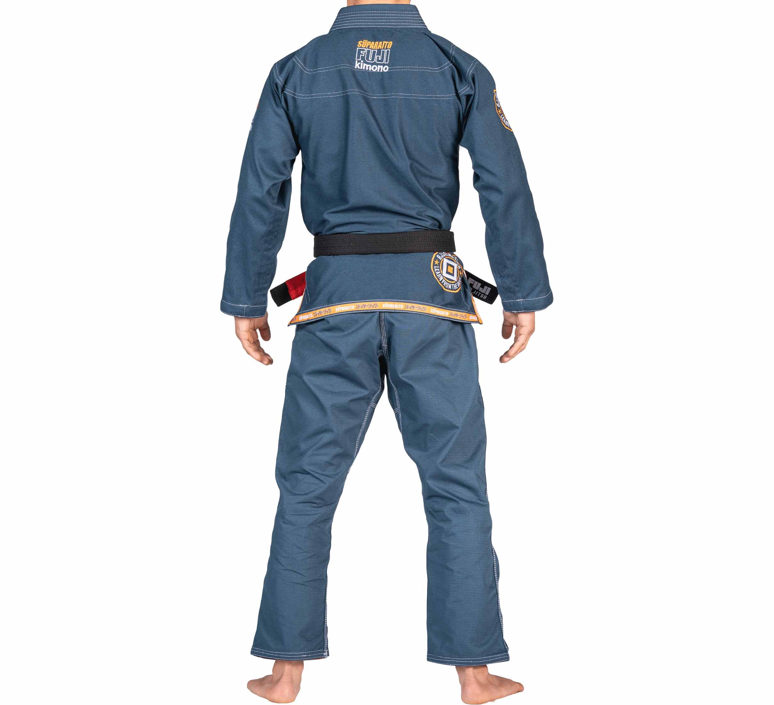 A barefoot person in a FUJI X BJJ Fanatics Blue Suparaito Gi and black belt stands with their back to the camera, arms relaxed, displaying various patches on the gi.